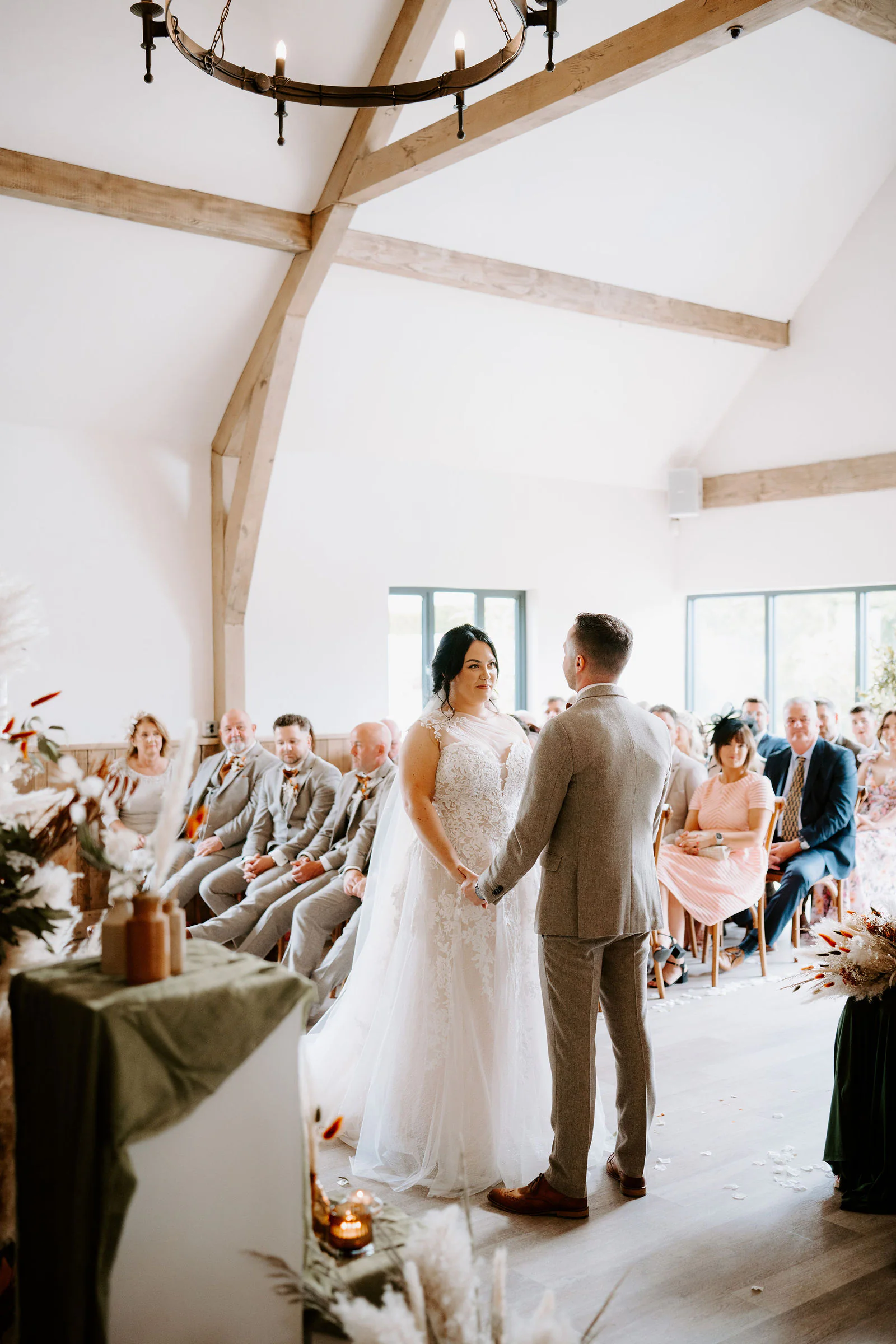 Hannah and Tom hold hands during their ceremony beneath pale timber beams at The River Barn, with guests seated either side