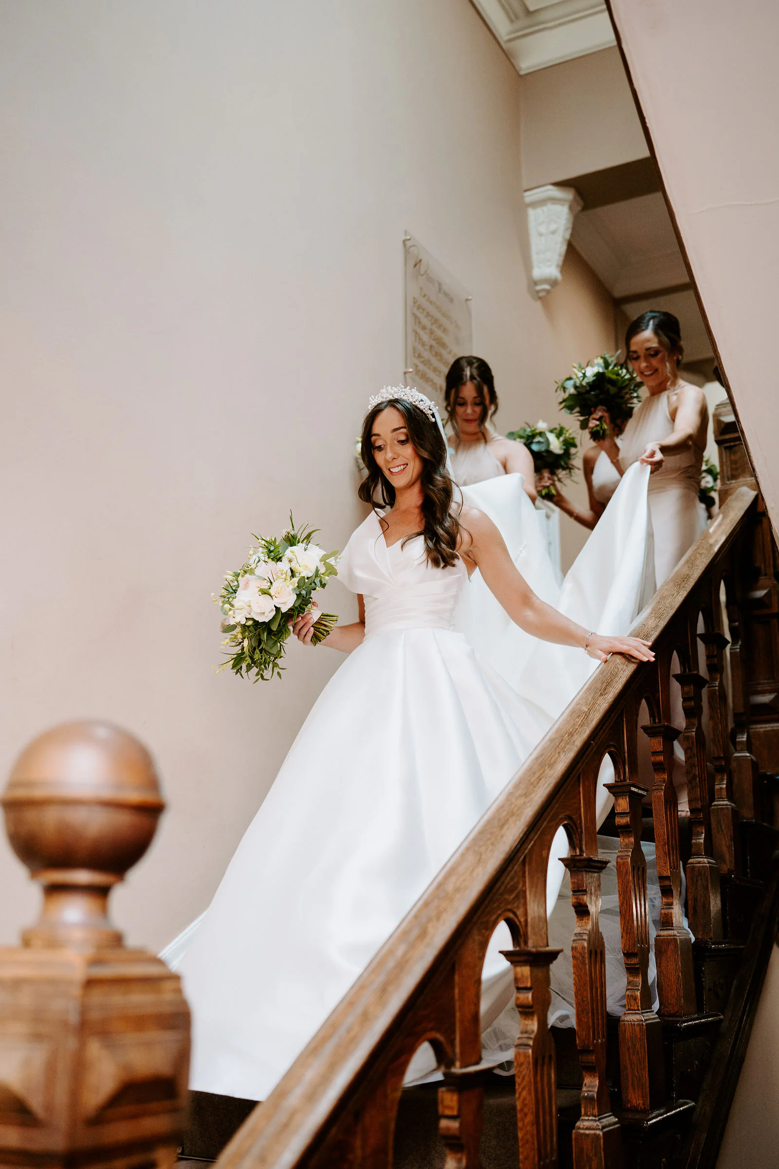 Natalie holding a bouquet comes down a wooden staircase with her bridesmaids behind her