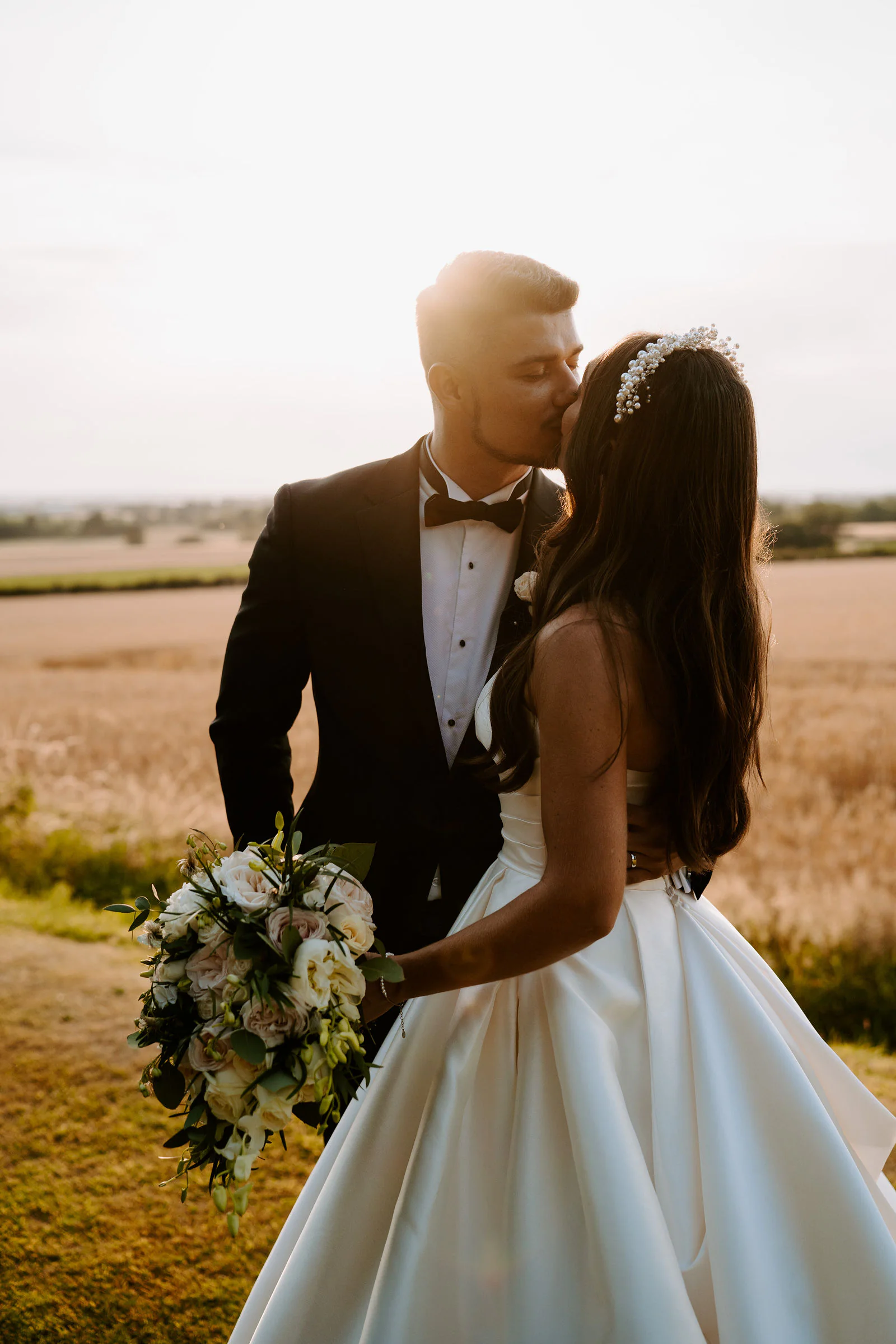 Natalie and Damian kiss in the evening light beside a field of wheat, Natalie holding a bouquet of white flowers
