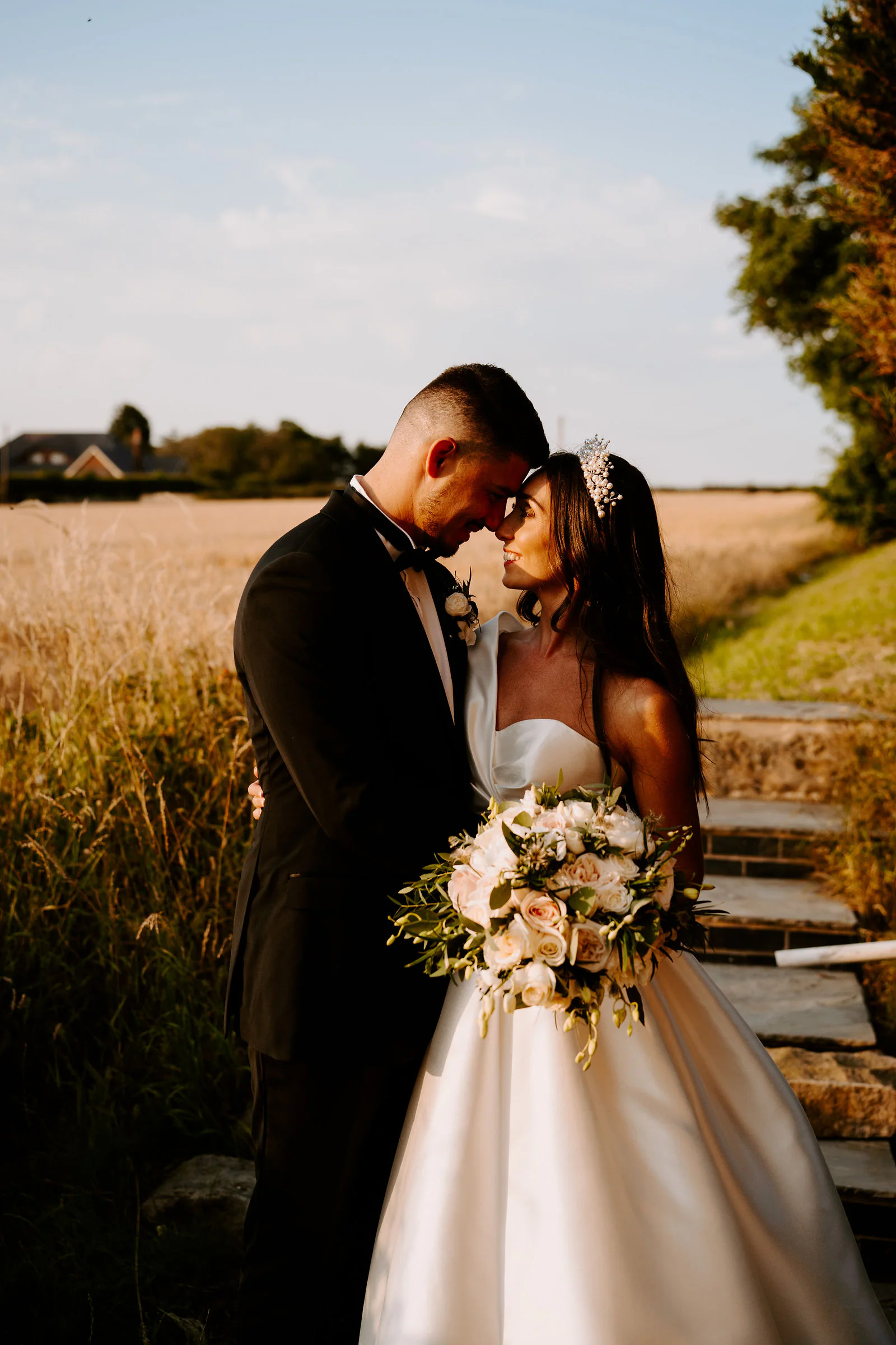 Natalie and Damian touch foreheads on stone steps in golden light with a field and a line of trees behind them