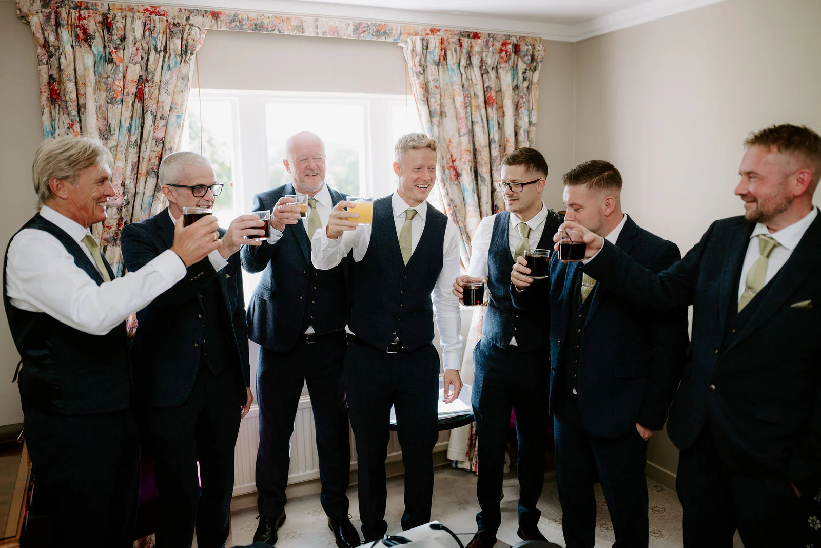 Will and his groomsmen raising their glasses in a bright room with floral curtains