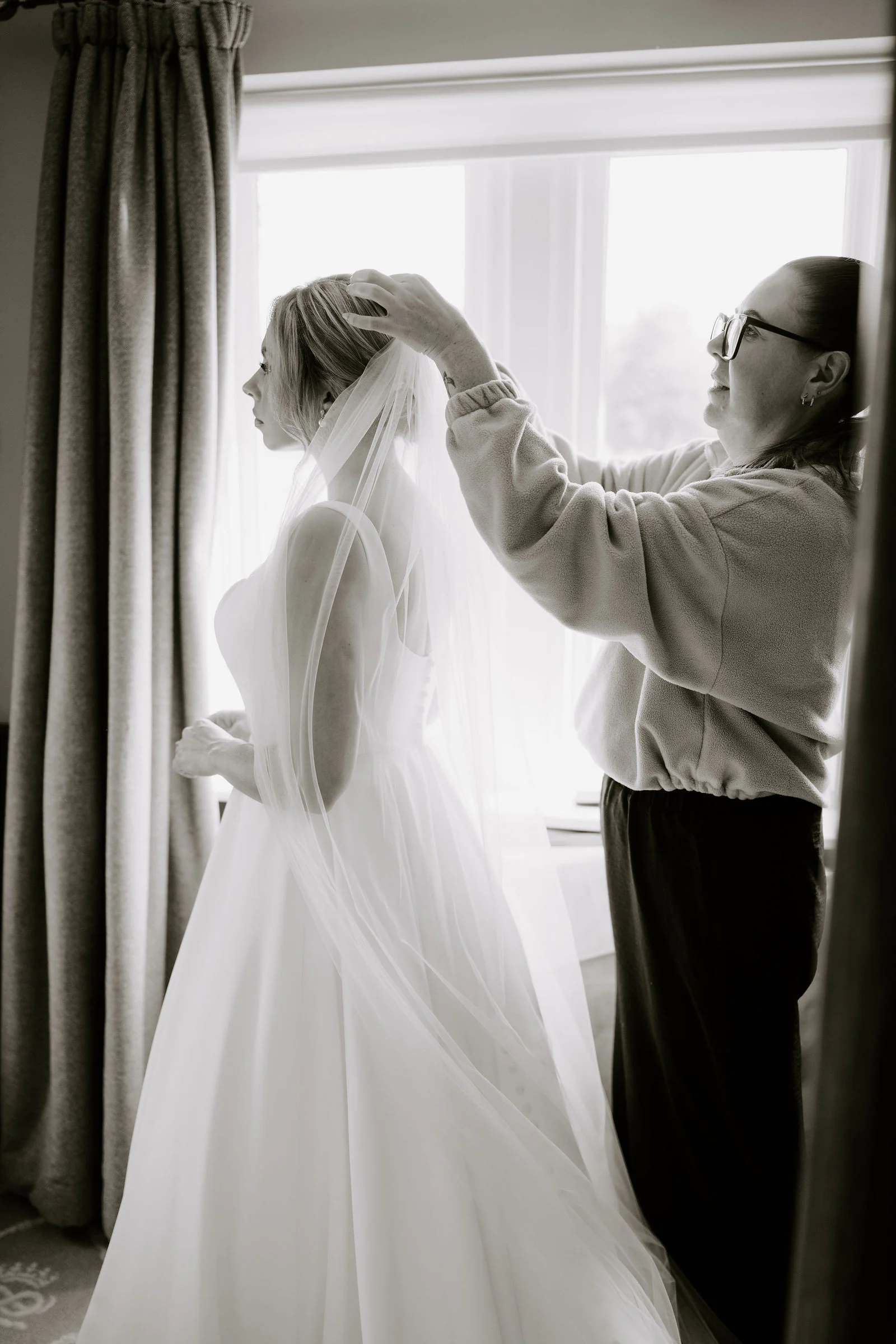 Amber in black and white at a window while a woman straightens her veil