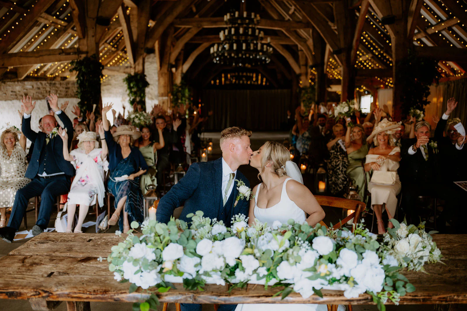 Amber and Will kissing over a table of white flowers inside the timbered Tithe Barn while guests cheer
