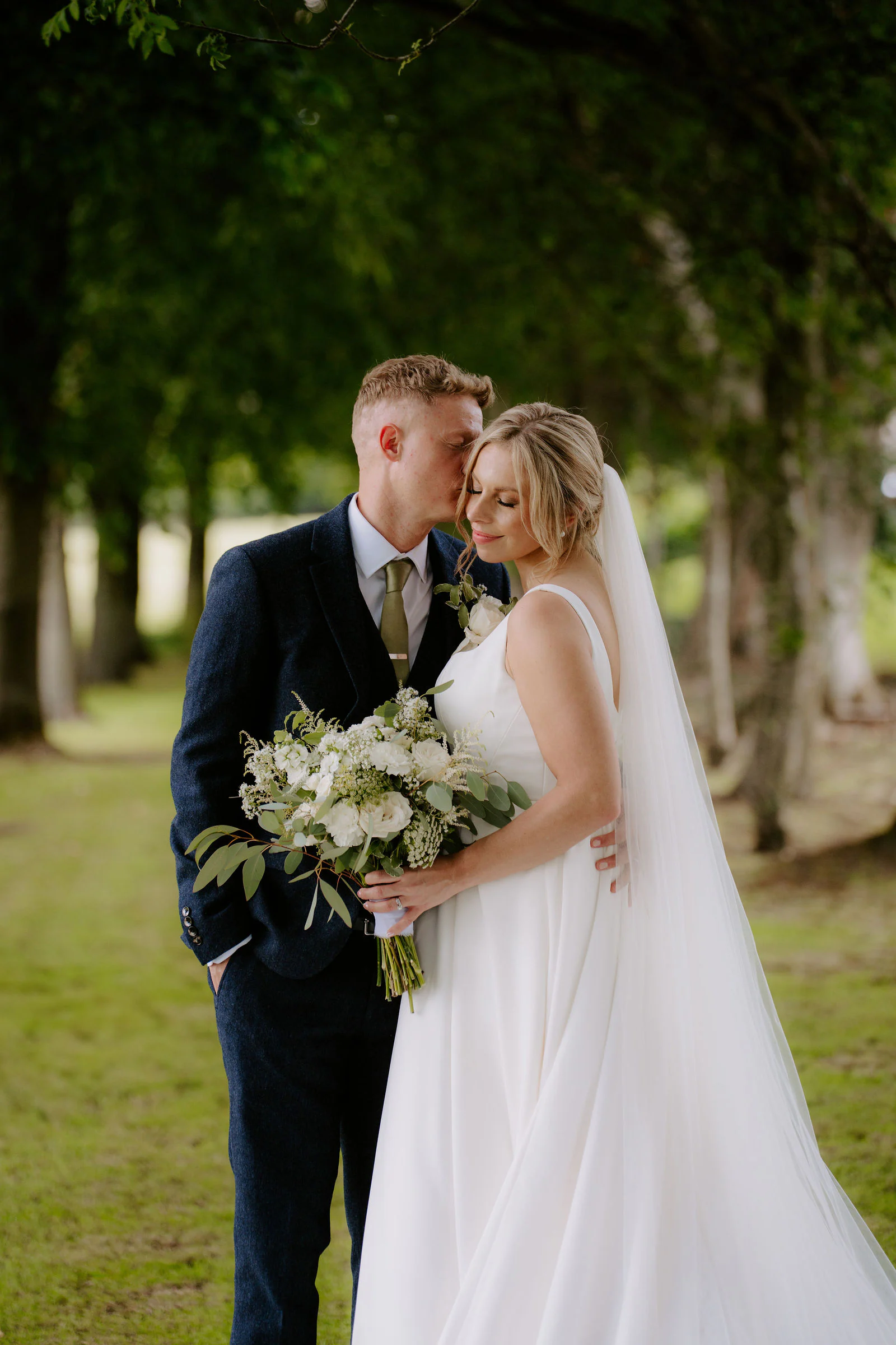 Amber and Will embracing on a lawn beneath tall trees with Amber’s bouquet held against her