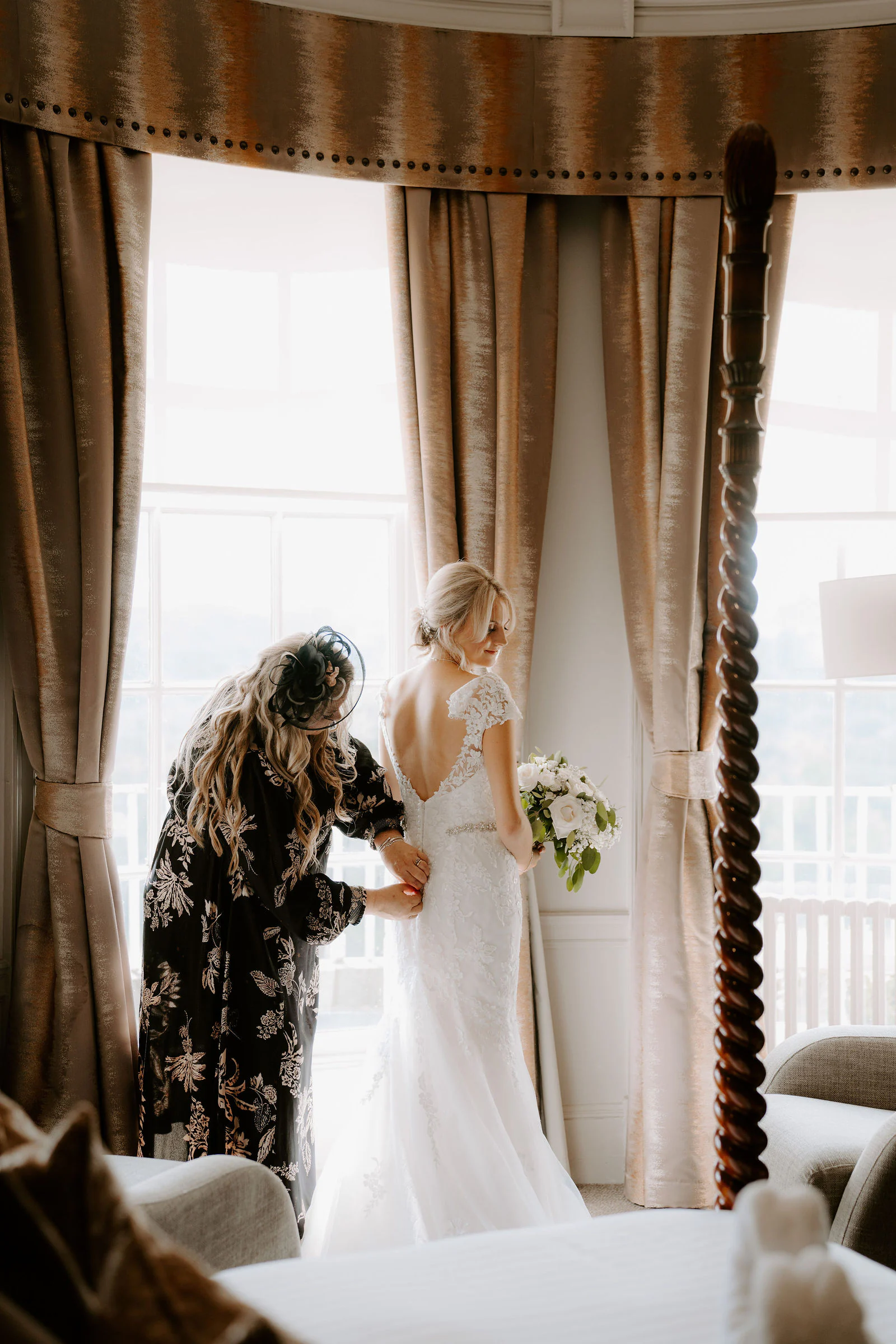 Brittany at a tall curtained window while a relative helps with the back of her wedding dress