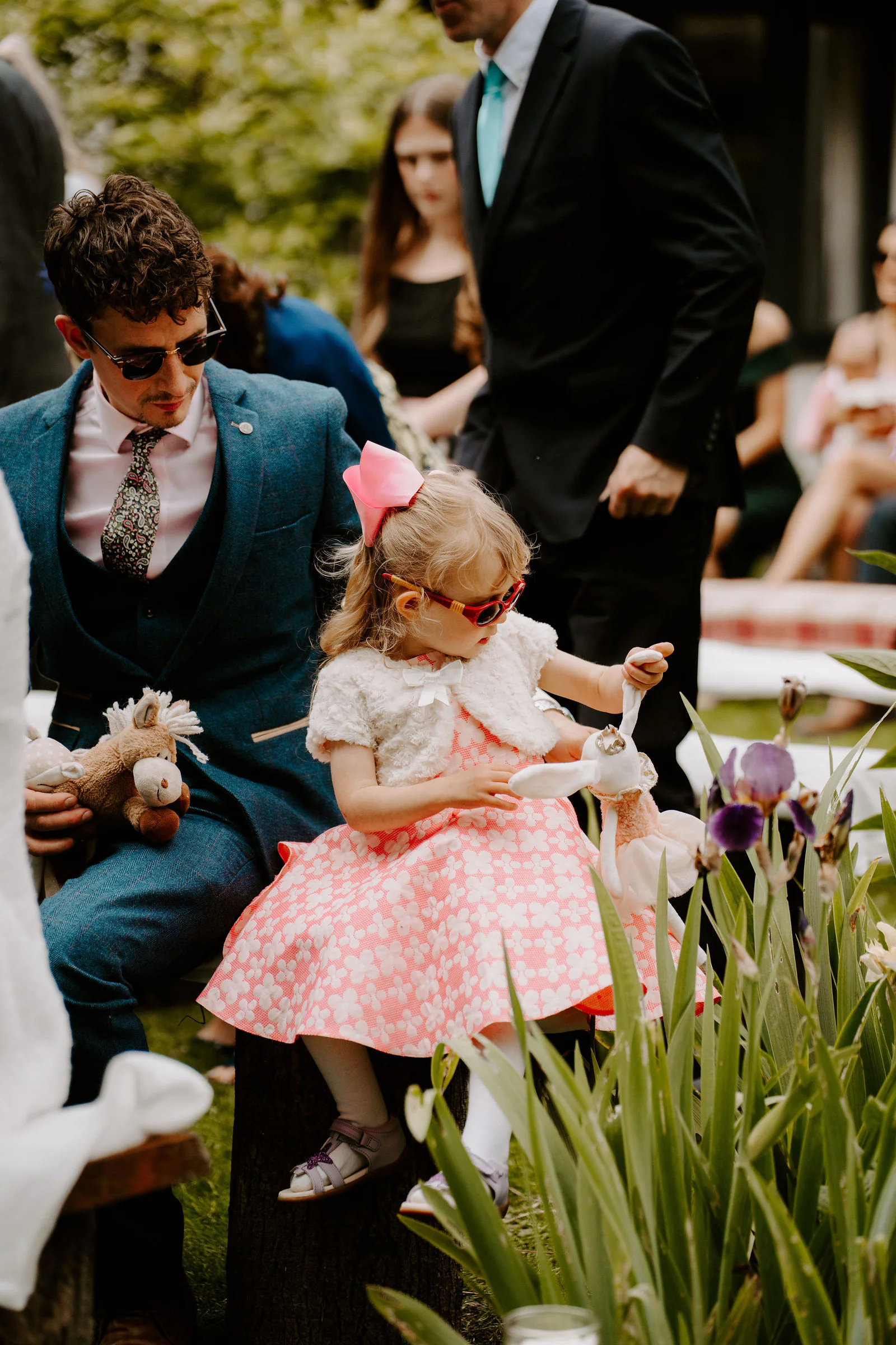 A young flower girl in a pink dress and sunglasses sits among purple irises at the outdoor ceremony as guests look on
