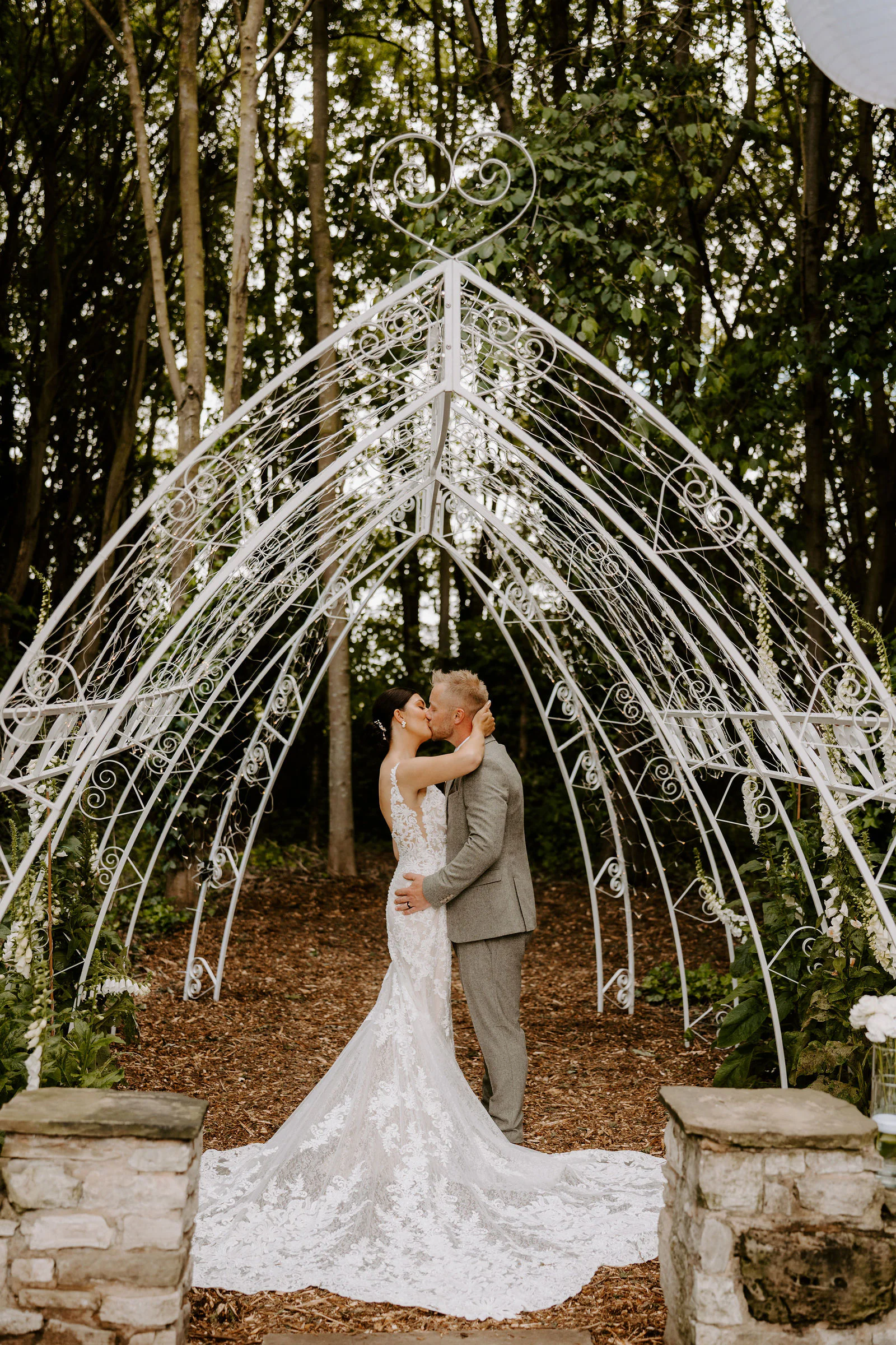 Philippa and James kiss beneath a white wrought-iron arch among trees, her lace train spread across the woodchip path