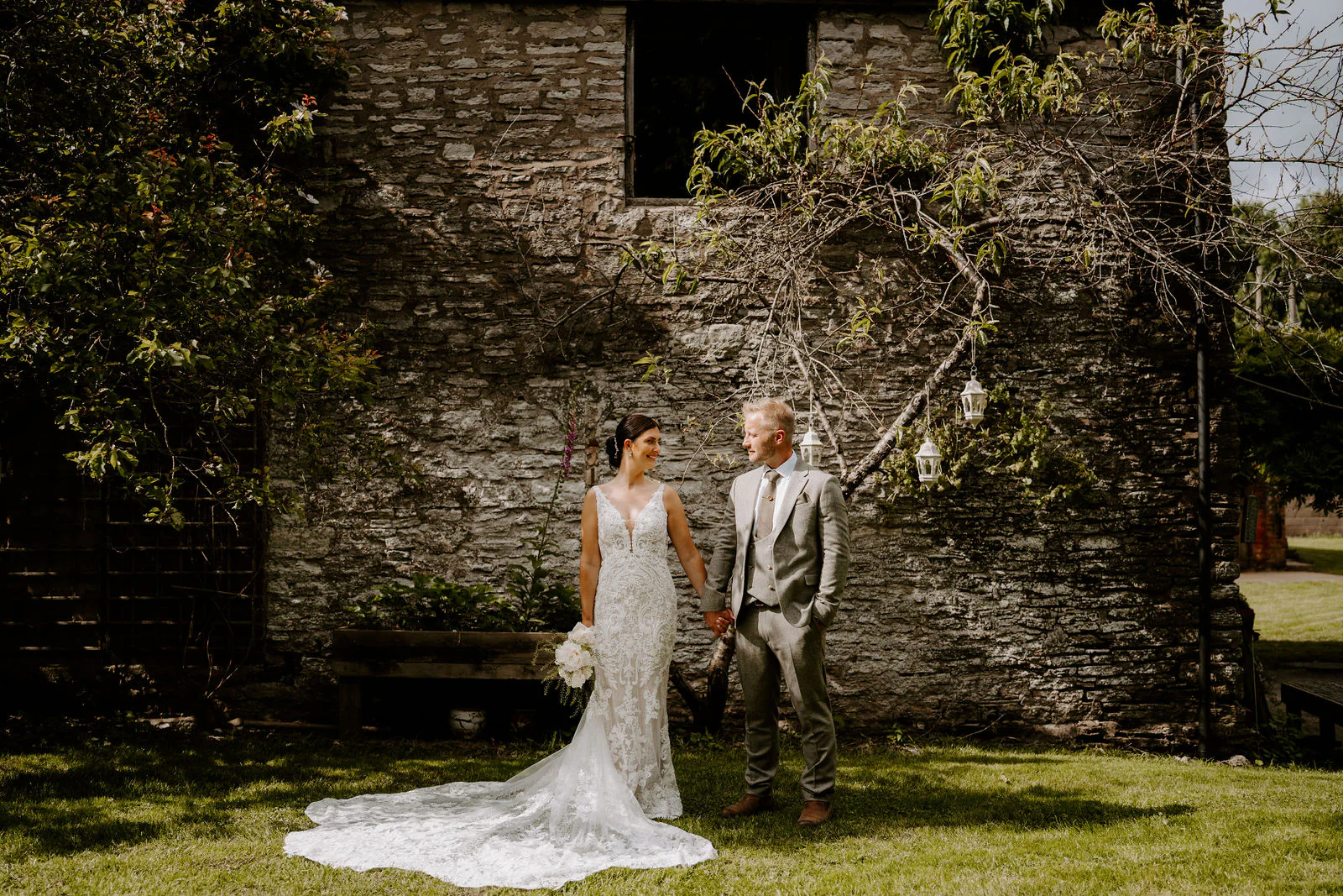 Philippa and James stand hand in hand on the lawn in front of an old ivy-covered stone building