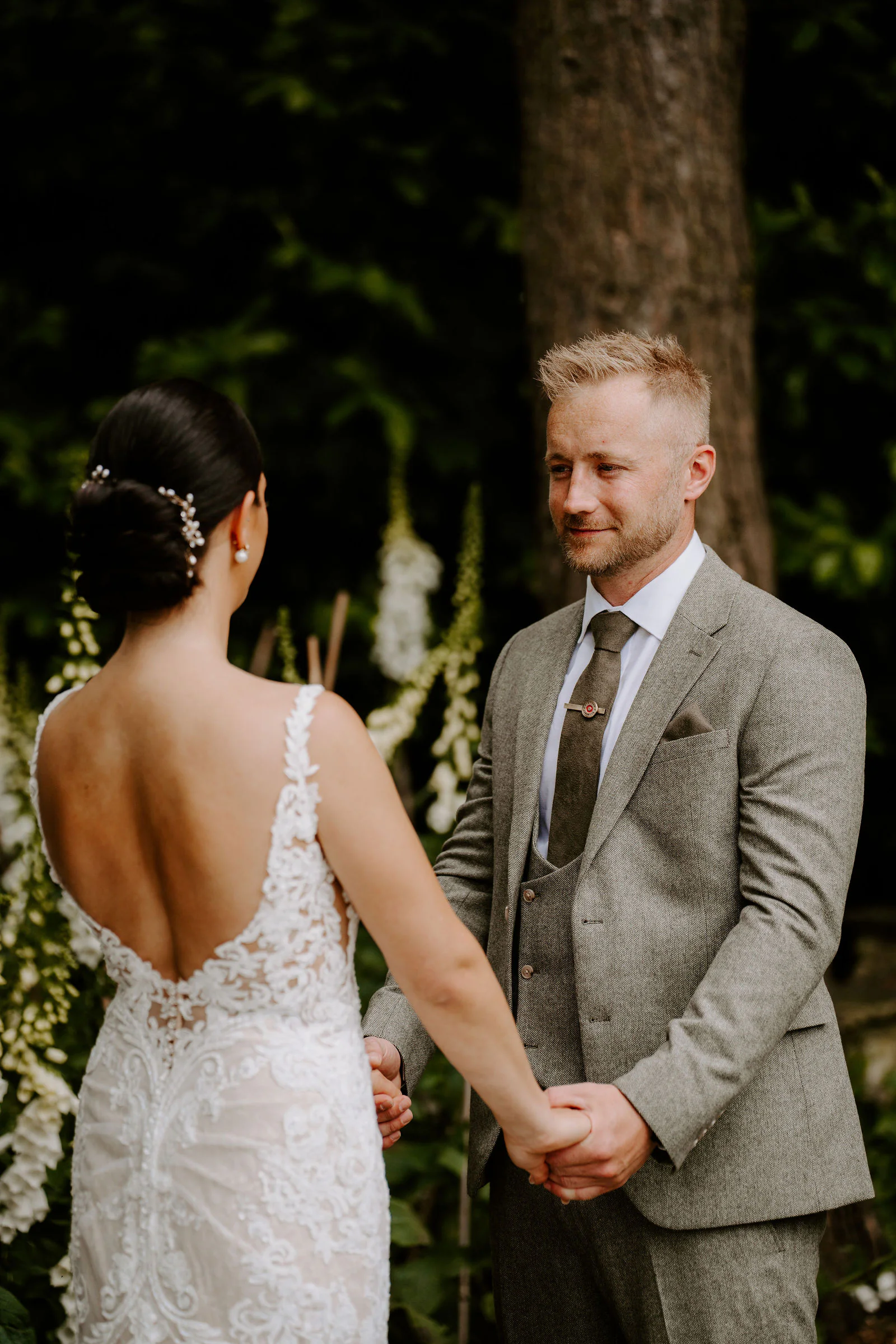 Philippa and James hold hands and smile at each other as they say their vows outdoors, tall white flowers behind them