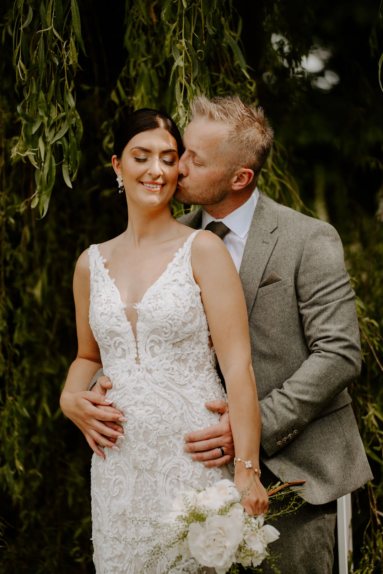 James kisses Philippa's cheek as she smiles with a bouquet of white flowers in her hand, willow branches hanging around them