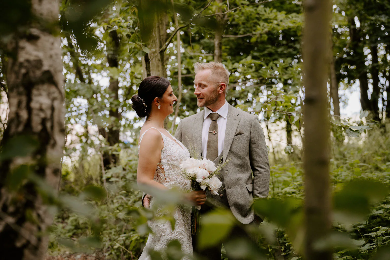 Philippa holding a bouquet of white flowers and James in a grey suit look at each other among trees