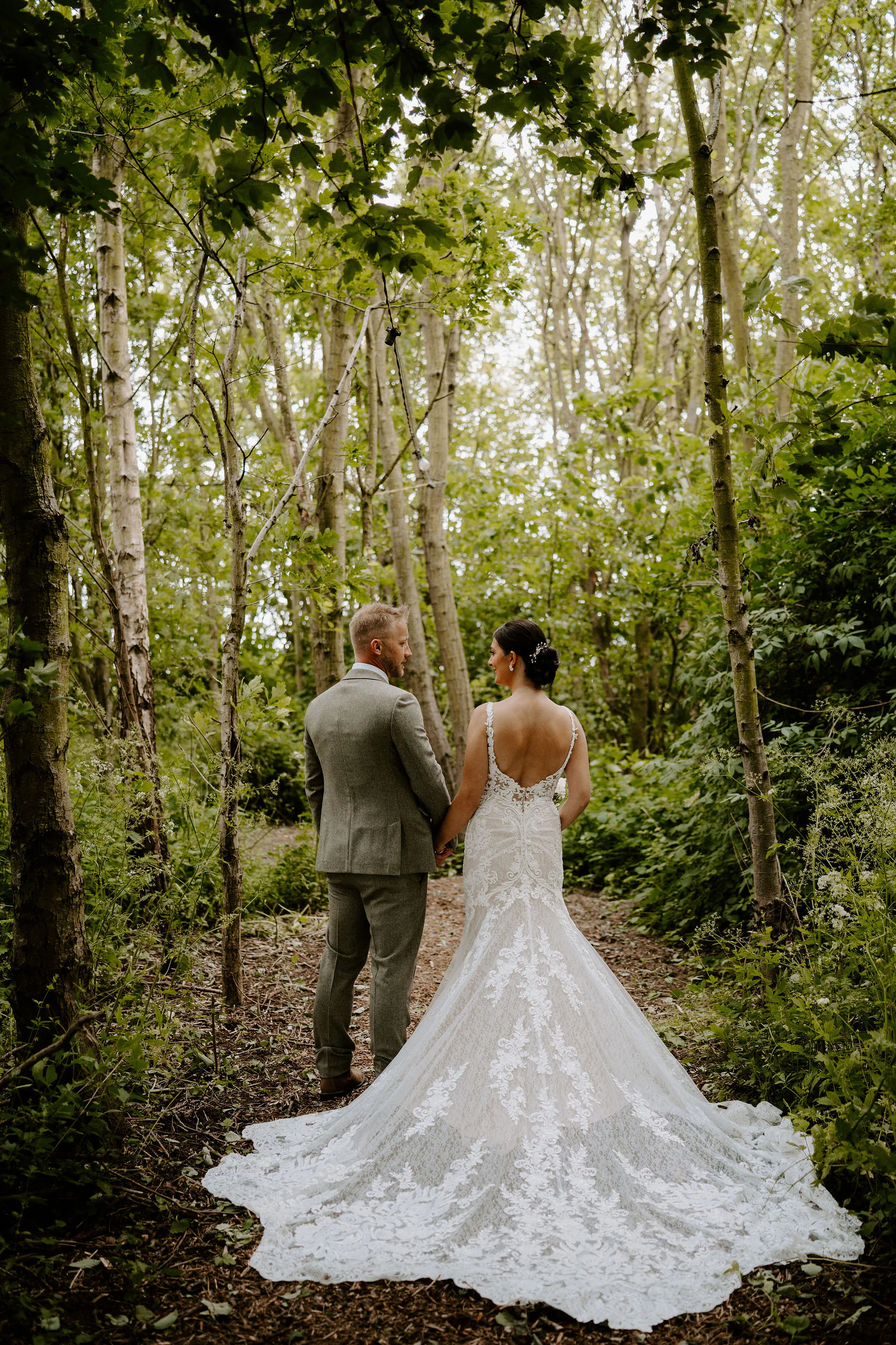 Philippa in a lace gown with a long train and James in a grey suit stand hand in hand on a woodland path beneath birch trees, seen from behind