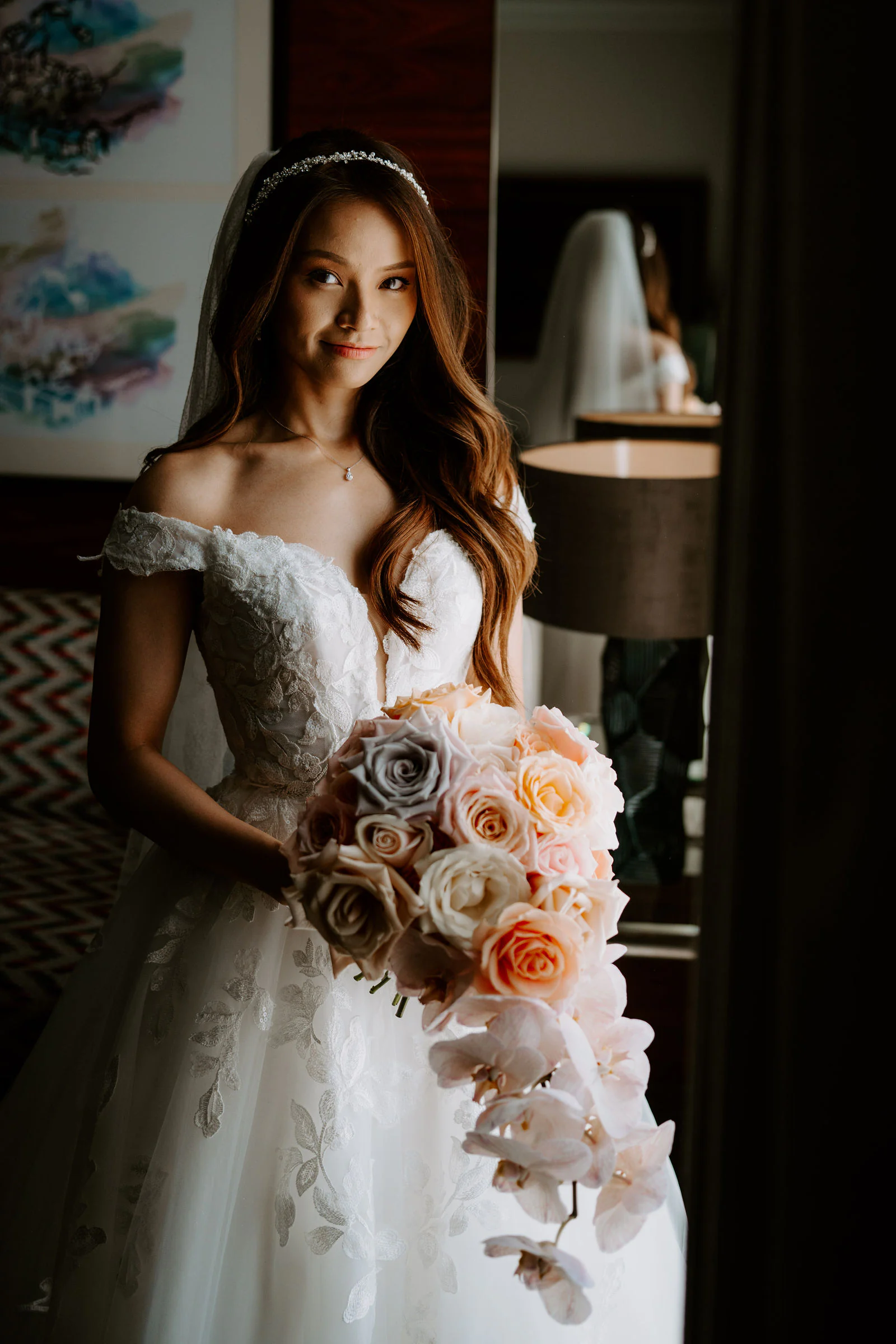 Sally in an off-the-shoulder wedding gown holding an apricot and cream bouquet in soft window light