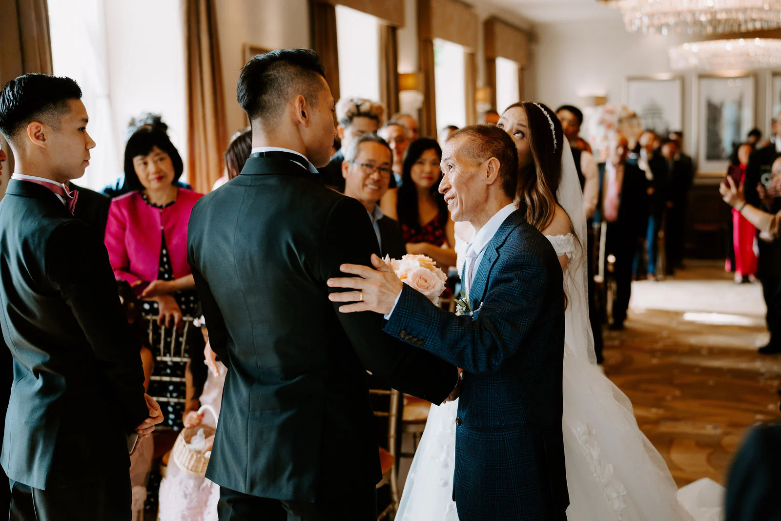 A man in a navy suit beside the bride greets the groom at the end of the aisle while guests look on at The Mansion in Leeds
