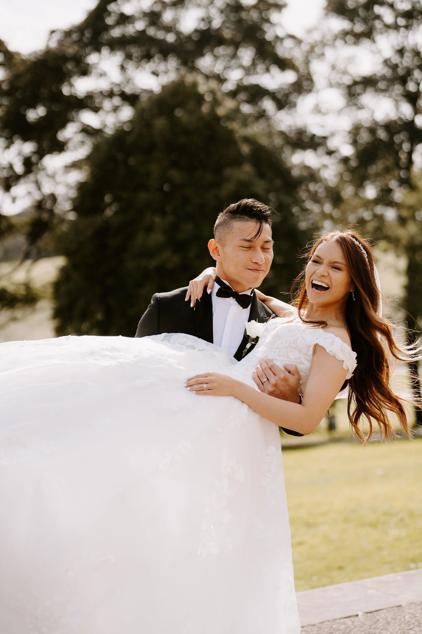 A groom in a black bow tie lifts his laughing bride in a wide white skirt, with large trees behind them