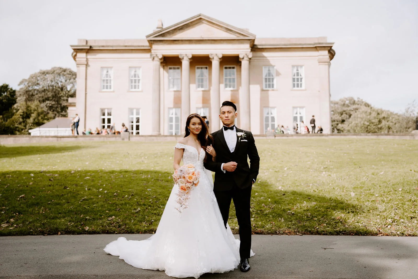 A bride holding a peach bouquet and a groom in black stand arm in arm on a drive in front of a white stone mansion with a tall columned portico, with guests on the terrace