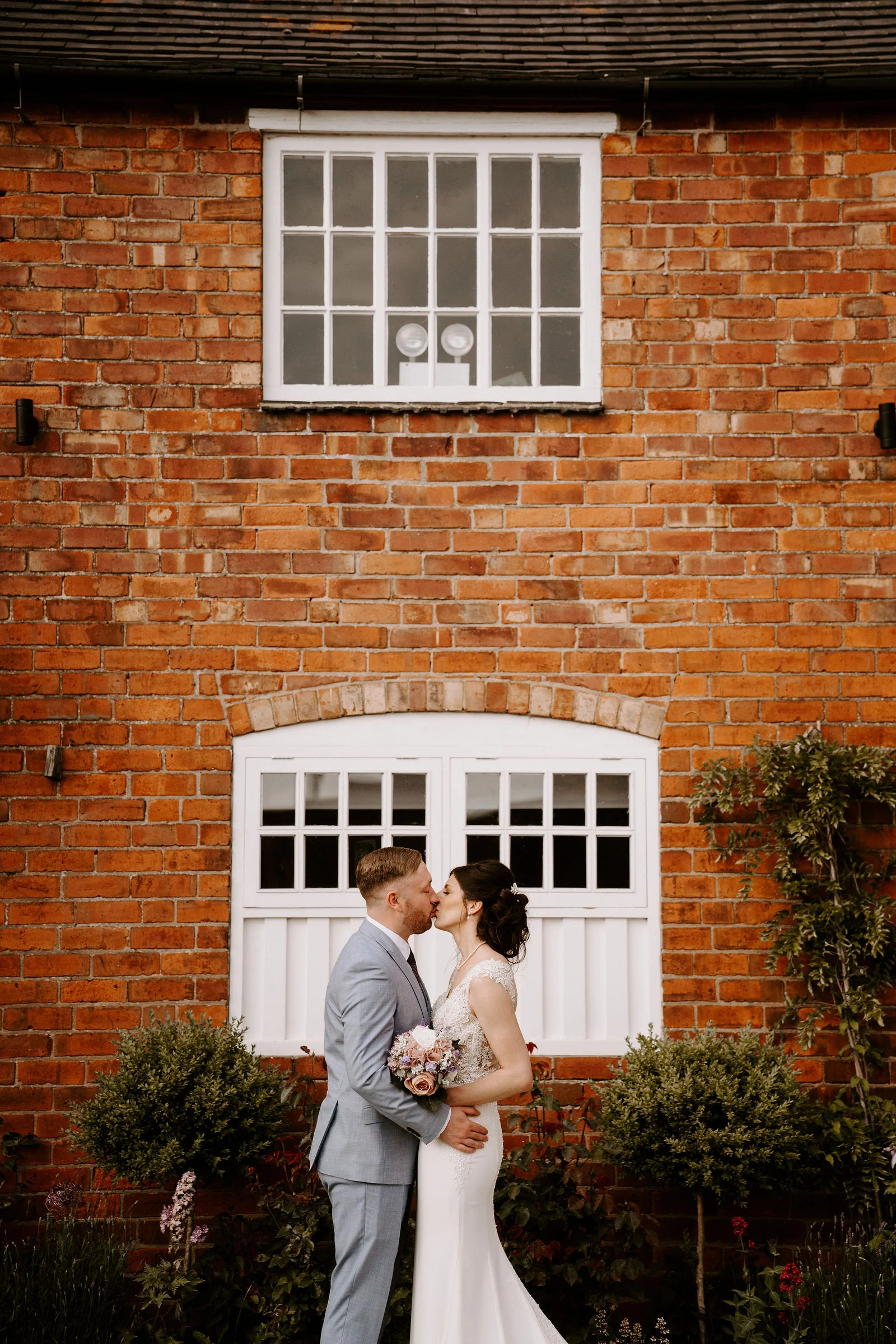 Kelly and Luke embracing beside the red-brick facade and white windows at Donington Park Farmhouse