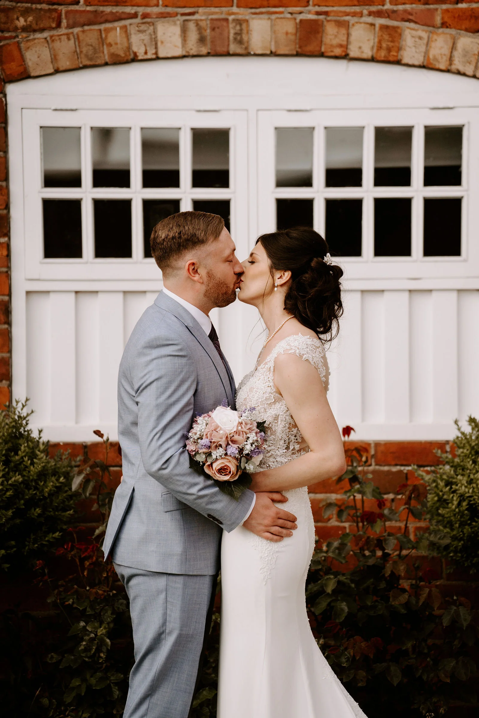 A groom in a light blue-grey suit kisses a bride holding a pink and lilac bouquet in front of a red-brick wall with large white arched doors