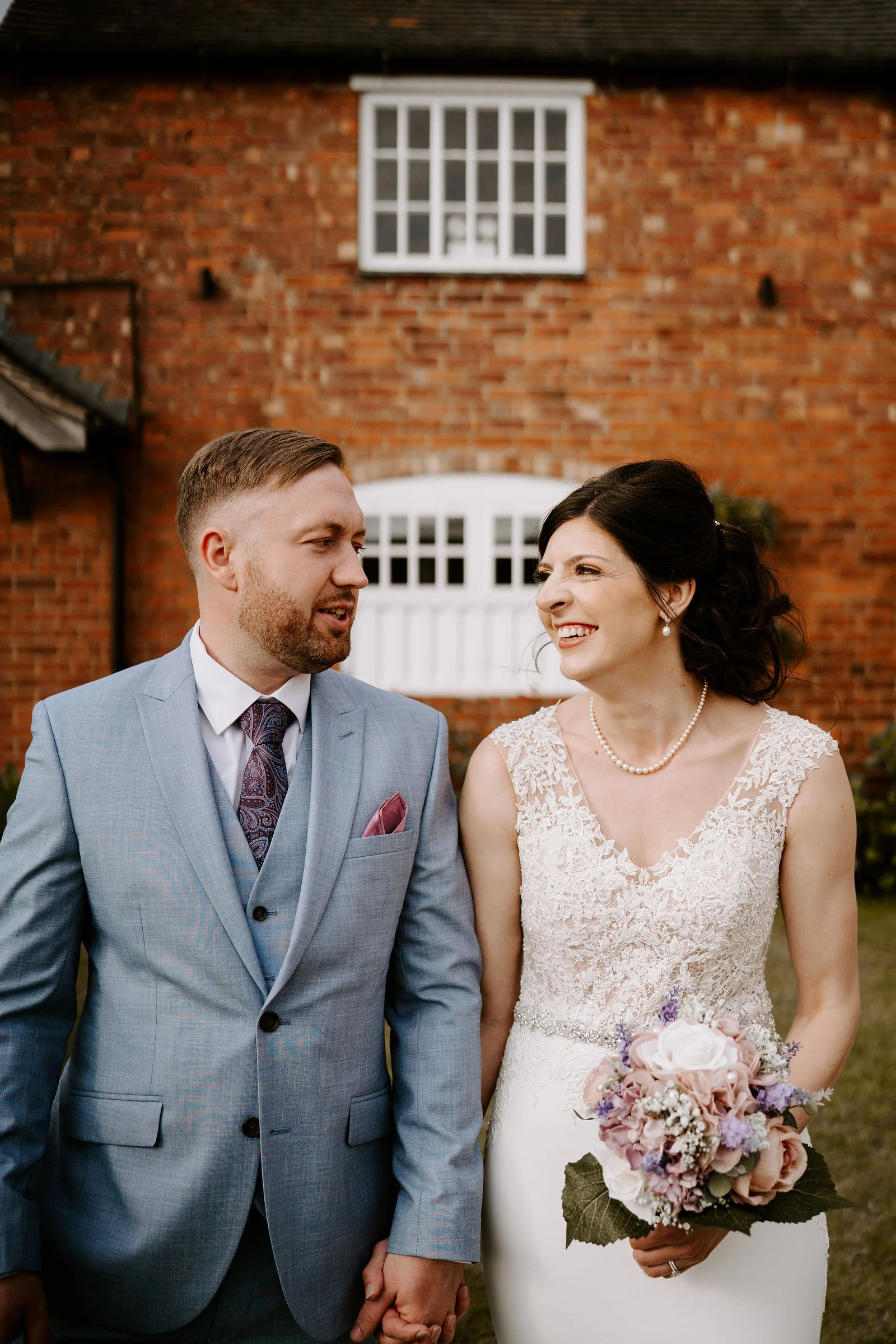 A smiling couple stand hand in hand in front of a red-brick farmhouse wall with white-framed windows, the groom in a light blue suit and purple tie, the bride in a lace bodice and pearl necklace with a pink and lilac bouquet