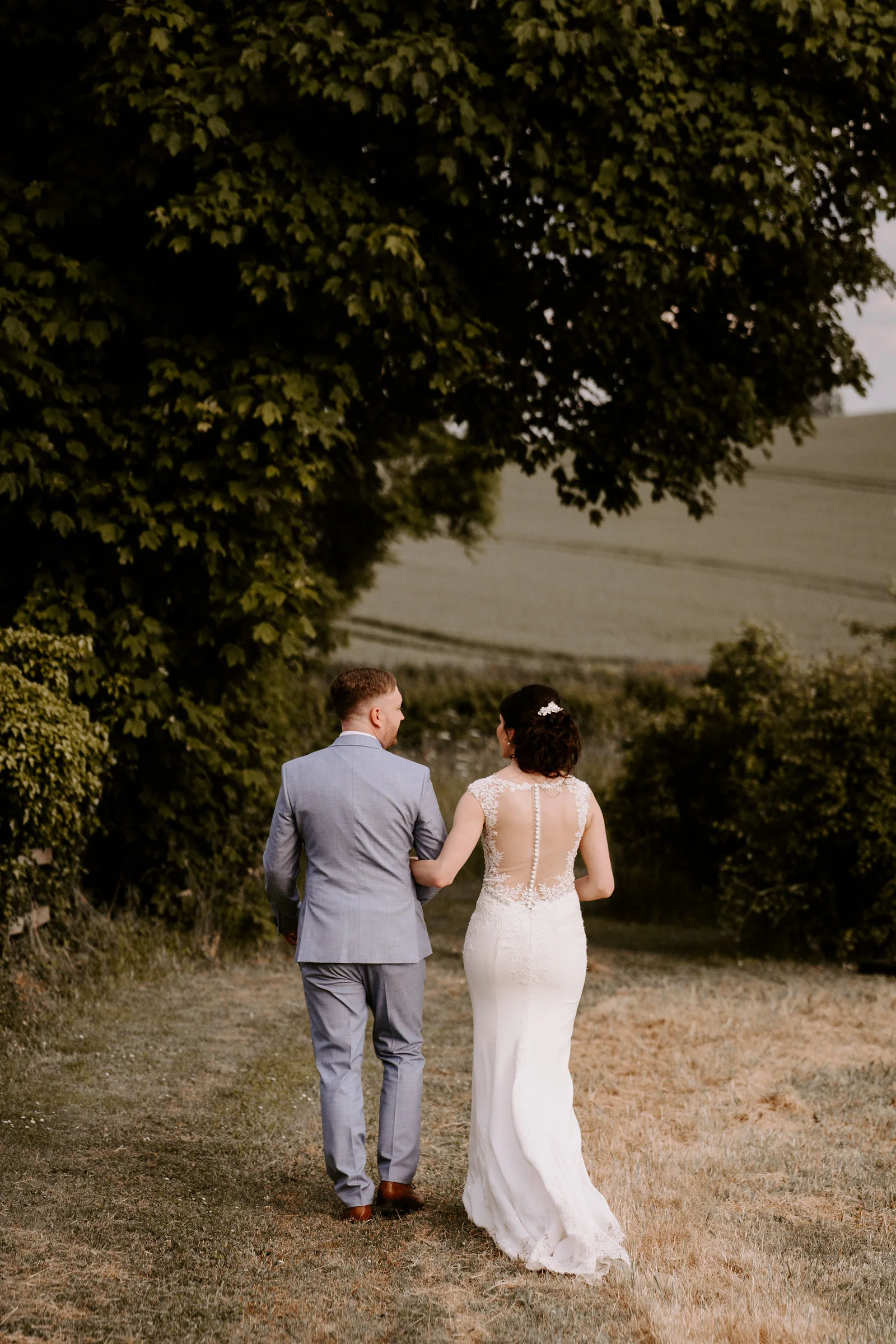 Seen from behind, a bride and a groom in a light blue-grey suit walk arm in arm along a mown track beside a hedge, under a large tree with a field rising behind