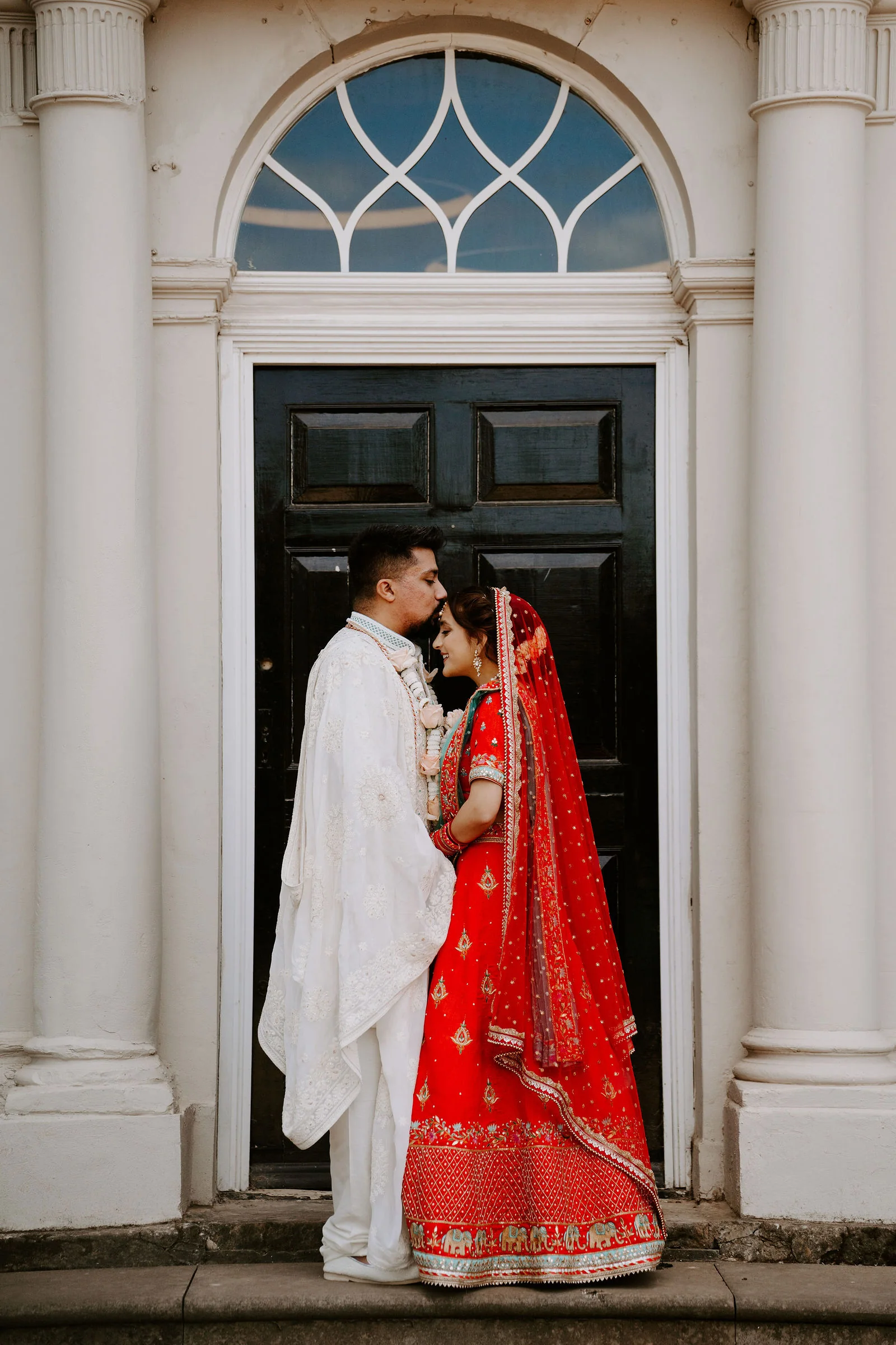 A groom in an ivory sherwani and a bride in a red and teal sari with a veil stand forehead to forehead in front of a black Georgian door with a fanlight and white columns