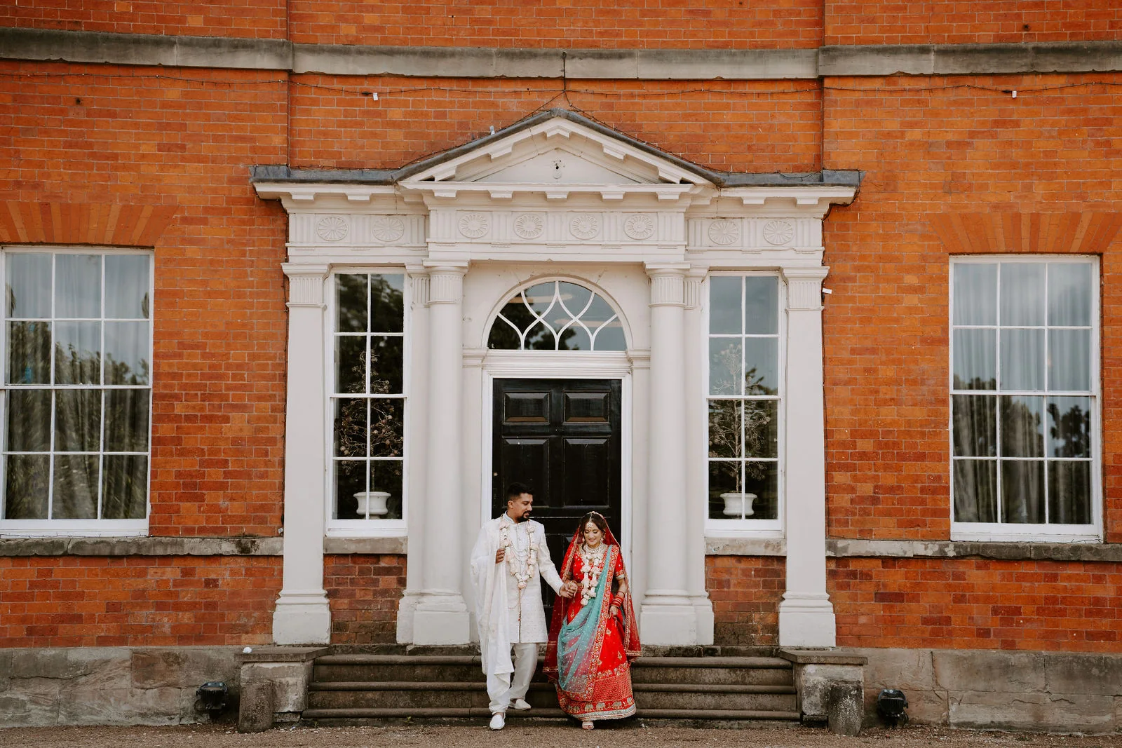 A wide view of a groom in an ivory sherwani and a bride in a red sari walking hand in hand down steps from the front door of a red-brick Georgian house with white columns and tall windows
