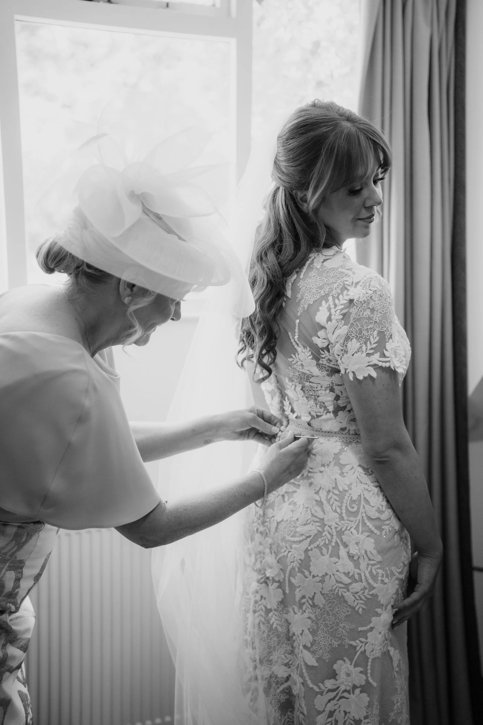 Amee having her wedding dress fastened beside a window, in black and white