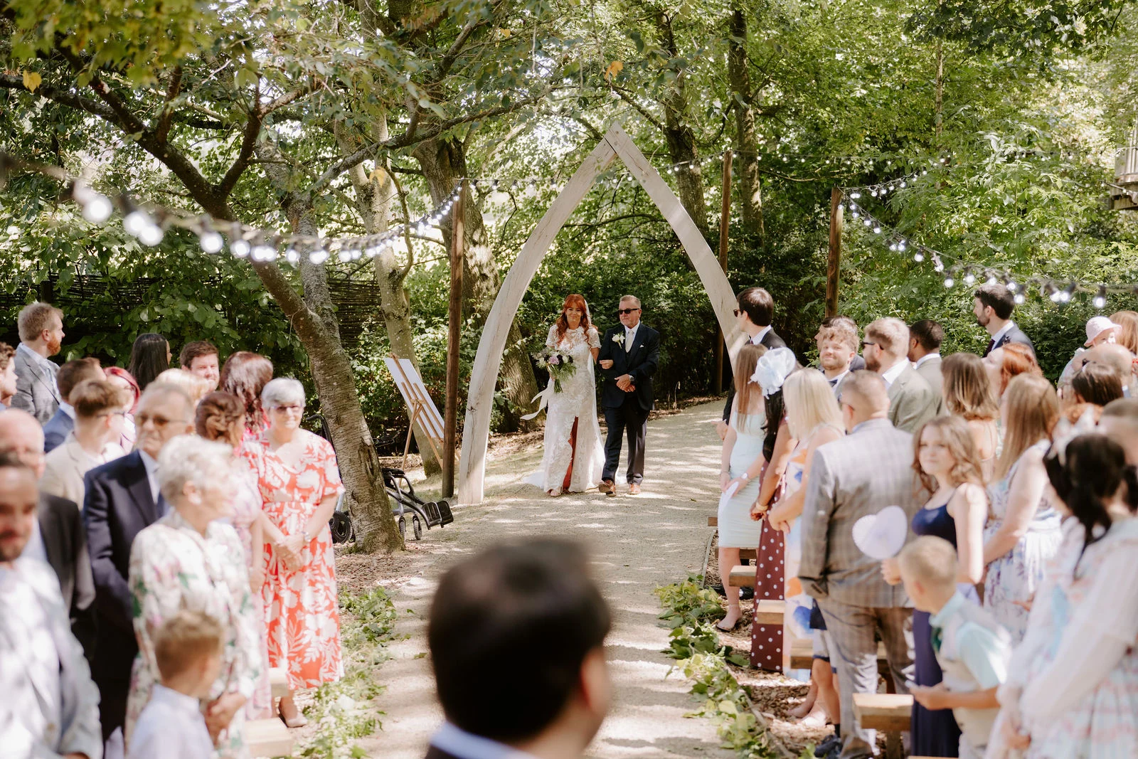 Amee walking towards Sean beneath a flower-covered woodland ceremony arch