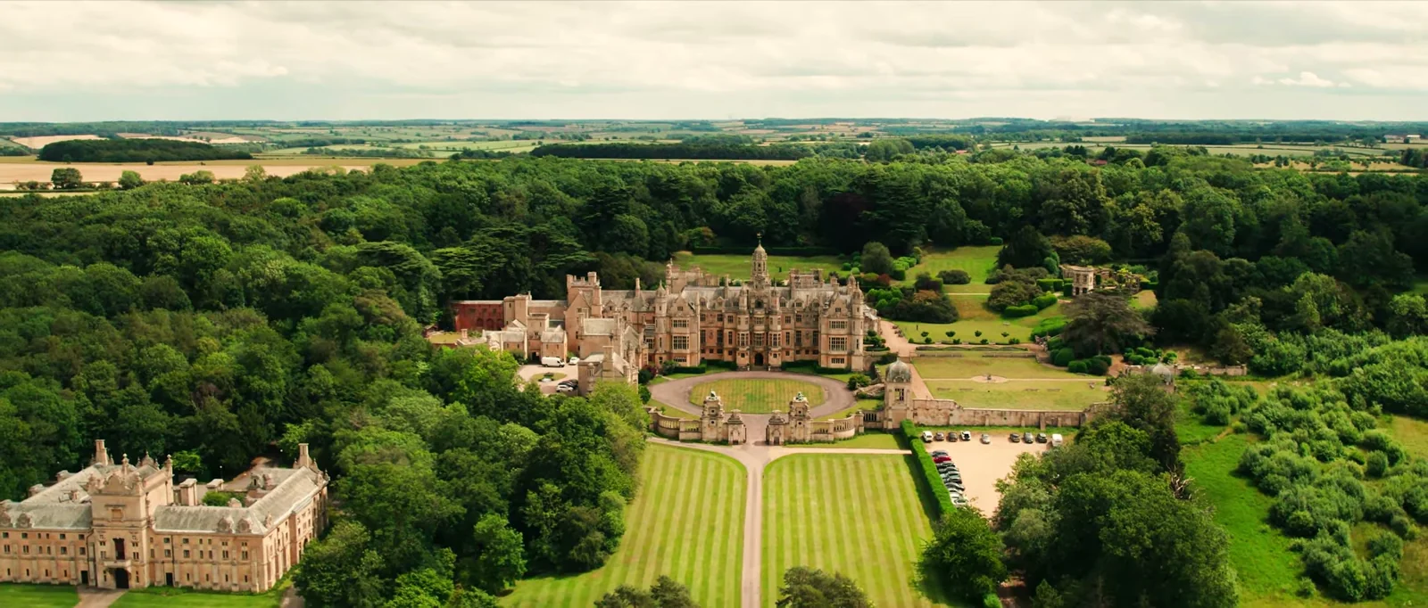 An aerial view of the stone manor with its round drive, striped lawns and formal gardens among woodland and fields