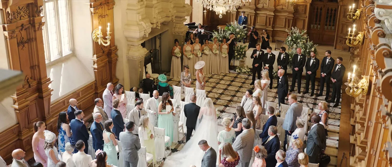 Seen from above, a bride in a long veil walks up the aisle of an oak-panelled hall on the arm of a man in a dark suit, with bridesmaids and groomsmen waiting at the front and guests standing on both sides