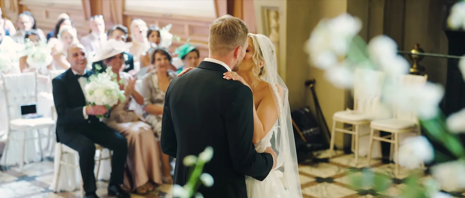 A couple kiss as guests behind them smile and clap, a man in black tie holding a bouquet on his lap