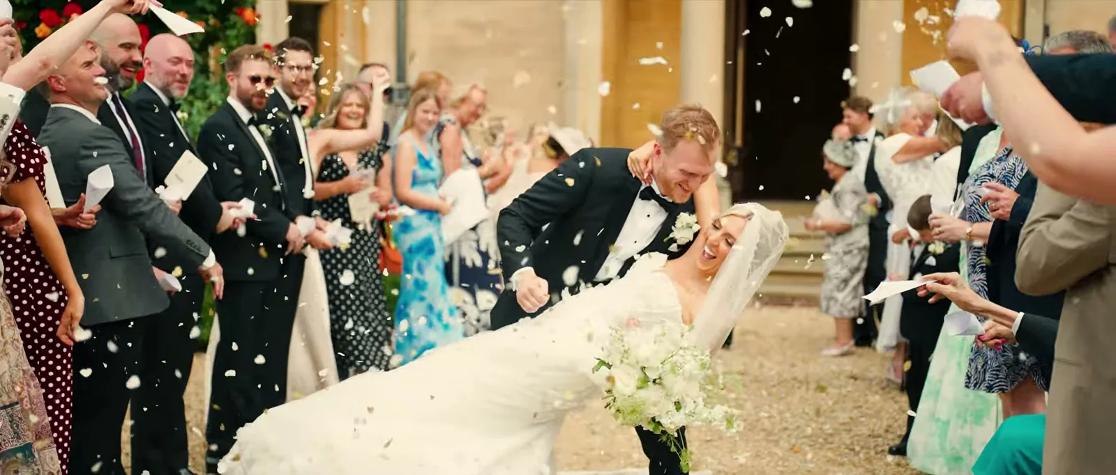 A groom dips his laughing bride as guests on both sides throw confetti outside the stone doorway of the manor