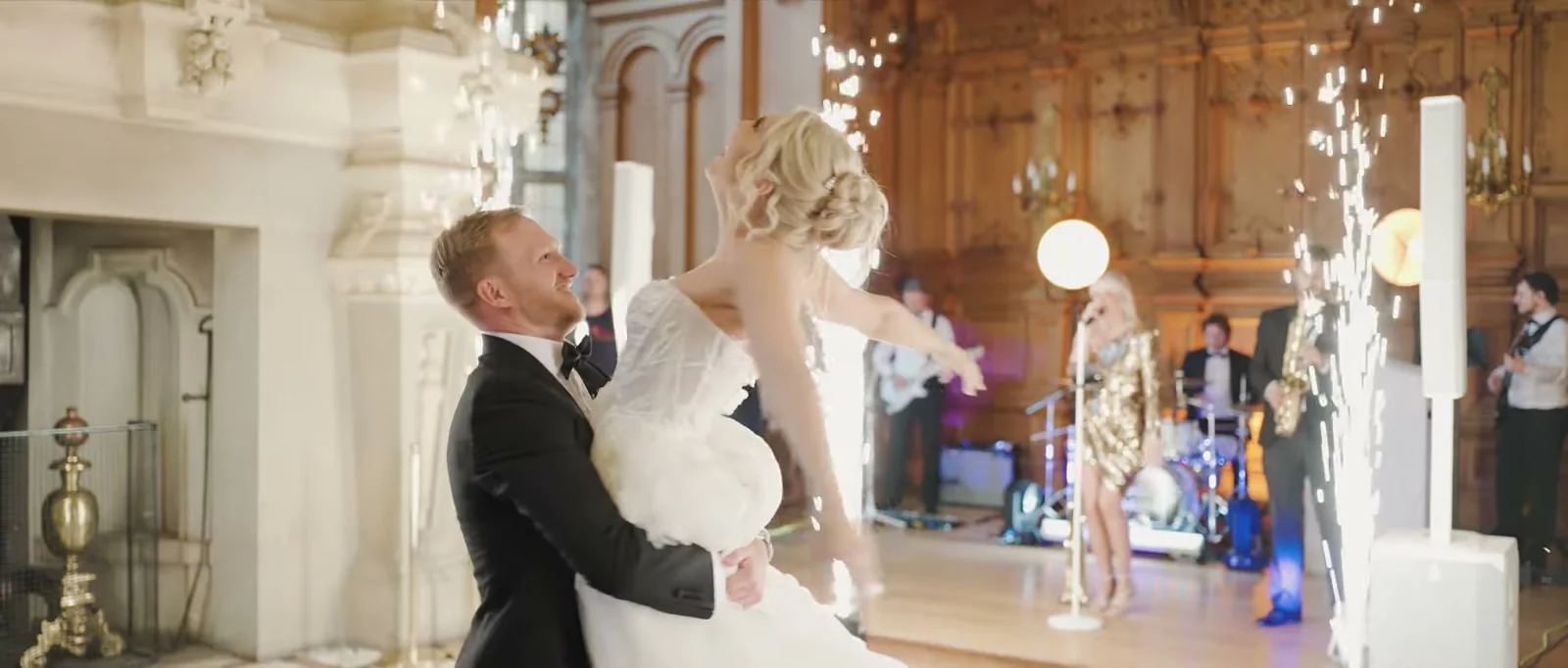 A groom lifts his bride as she leans back with her arms out, between two columns of sparklers on the dance floor with a band behind