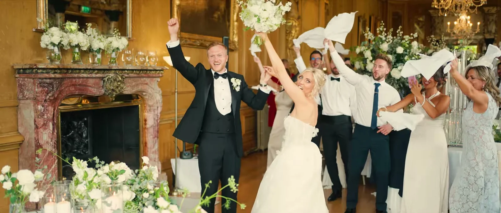 A groom and a bride with her bouquet raised cheer in a gold-panelled room with a pink marble fireplace while friends wave white napkins behind them