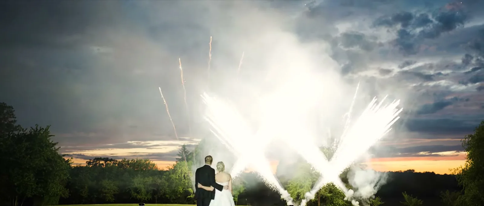 A couple stand arm in arm in silhouette on a lawn watching fireworks against an evening sky