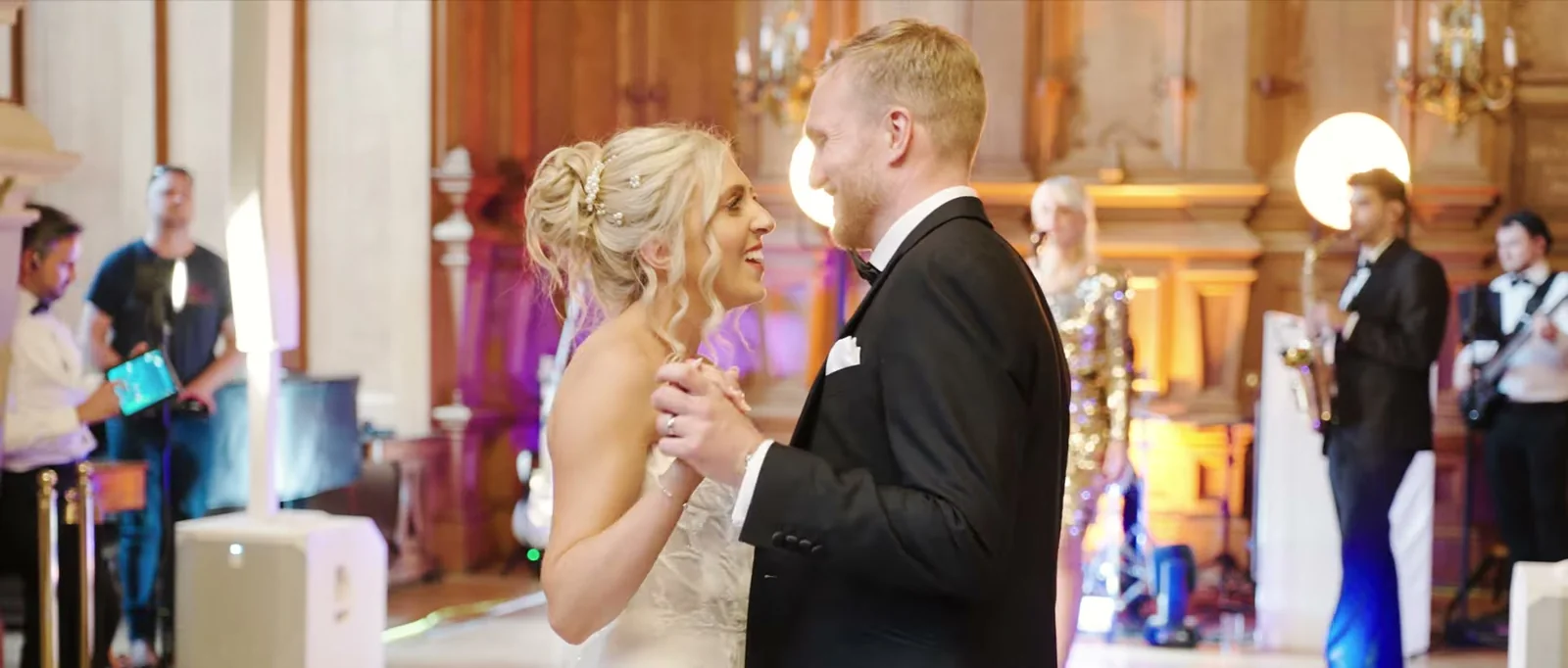A groom and a bride laugh as they hold hands for their first dance in a panelled hall, with a live band behind them