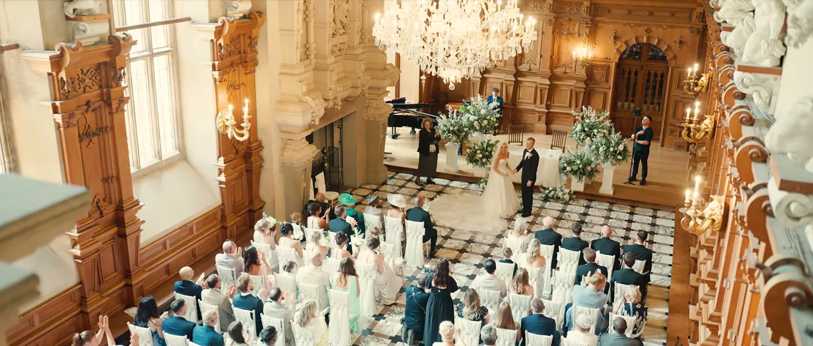Seen from above, a bride and groom stand hand in hand at the front of a panelled hall with a crystal chandelier, tall white flowers and a piano, while seated guests clap