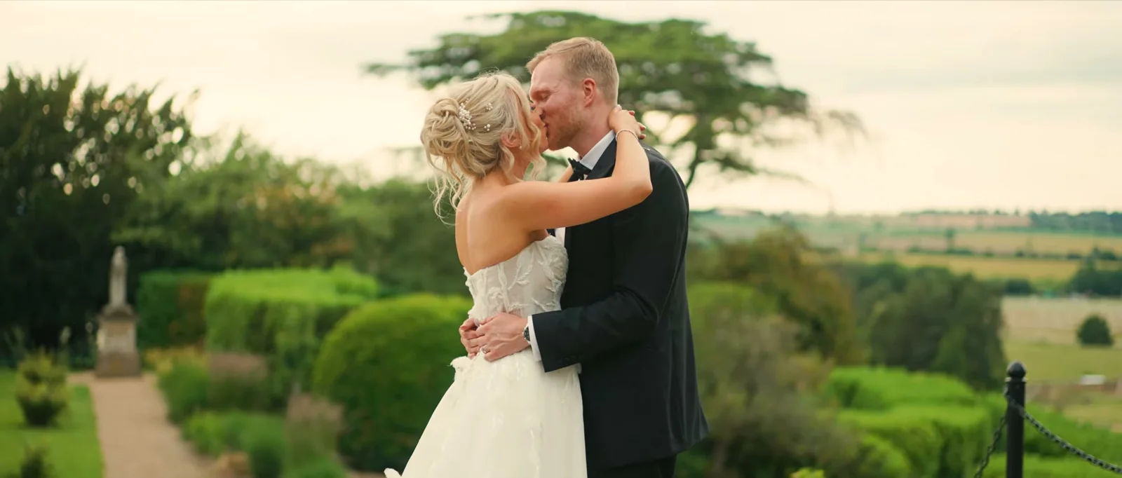 A bride and groom kiss at the edge of a garden terrace with clipped hedges, a statue and open countryside behind them