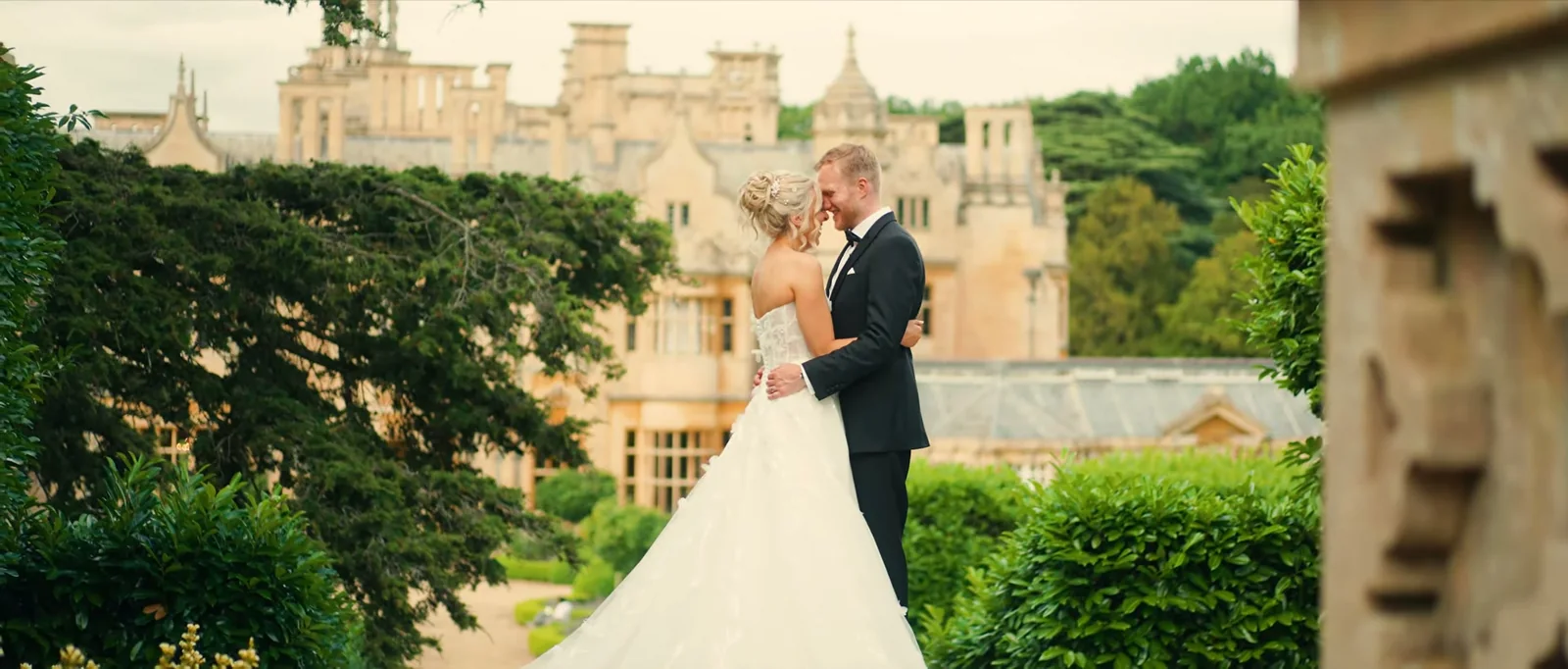 A bride in a strapless gown and a groom in a black tuxedo stand forehead to forehead among trees and clipped hedges with the stone front of Harlaxton Manor behind them