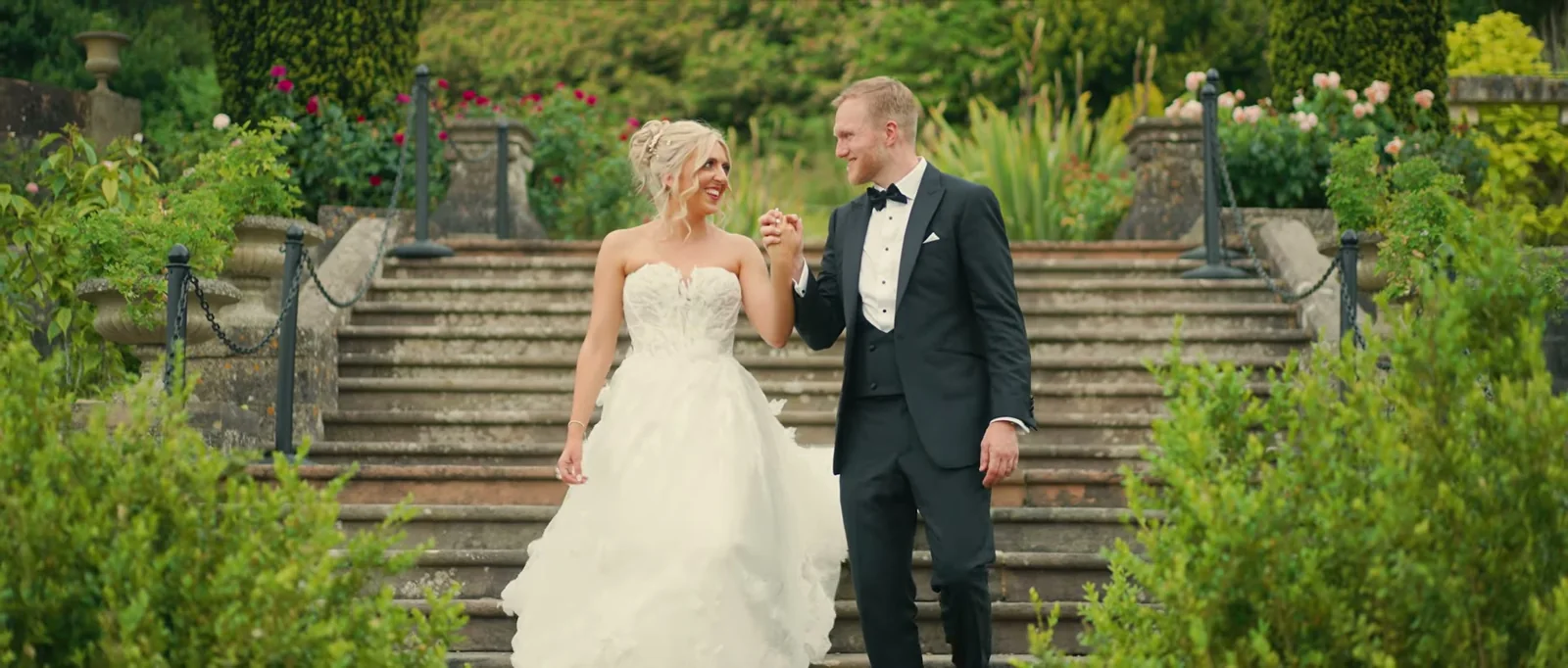 A bride and groom walk hand in hand down a flight of worn stone steps among roses and clipped yew, smiling at each other
