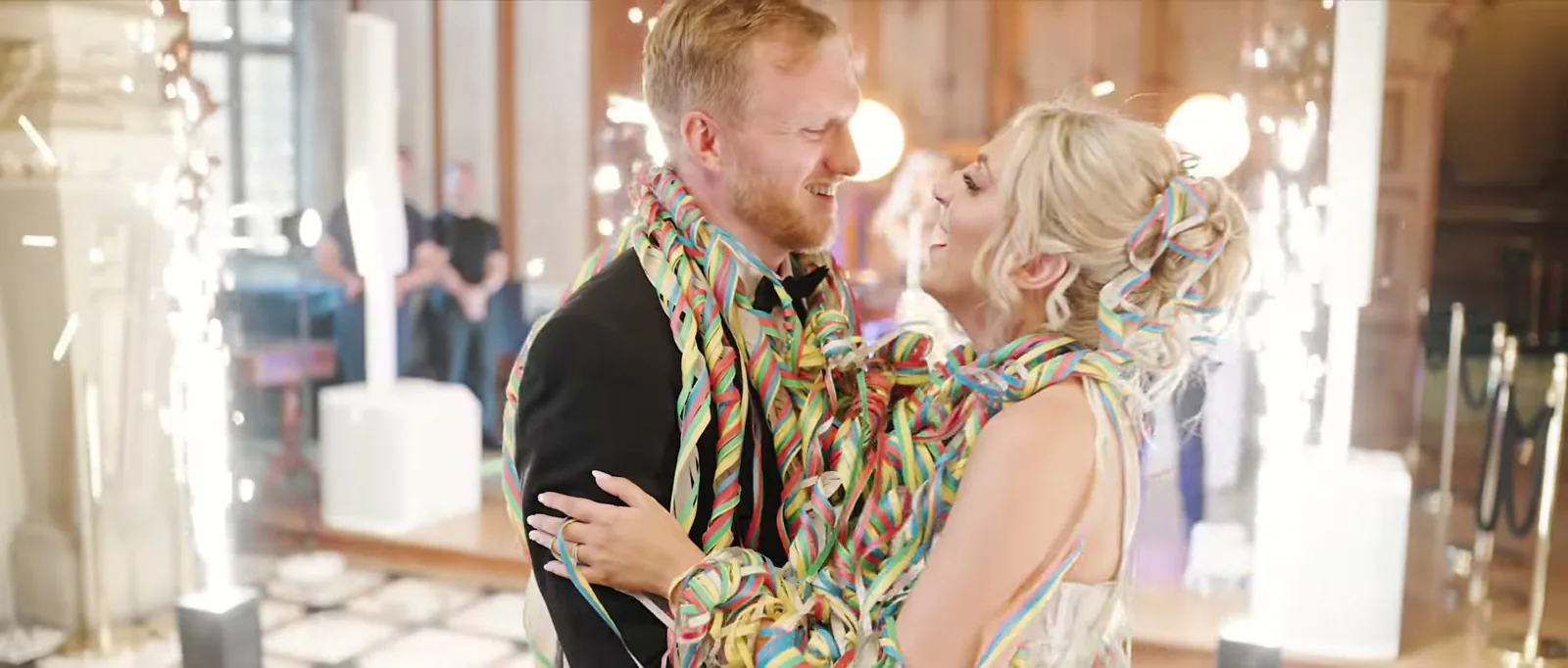 A groom and a bride laugh at each other with colourful streamers wrapped around both of them and sparklers behind