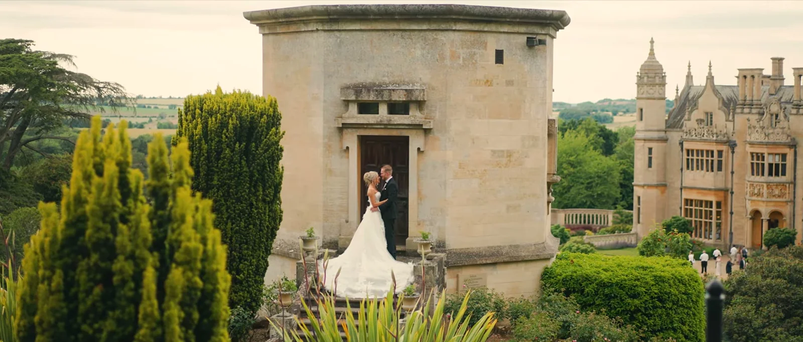 A couple embrace in the doorway of a round stone building in the gardens, with the manor to one side and fields beyond