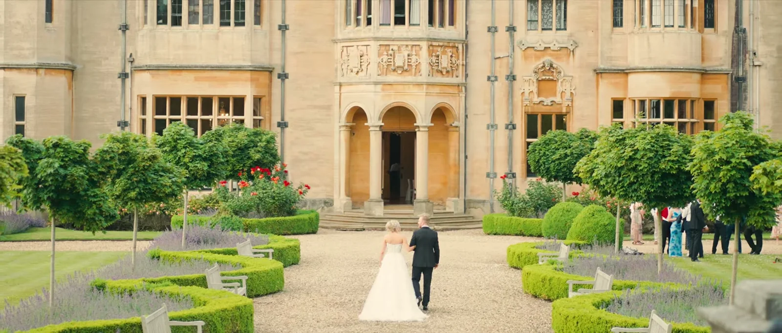A bride and groom walk arm in arm across gravel towards the arched doorway of the manor between clipped hedges and purple flowers