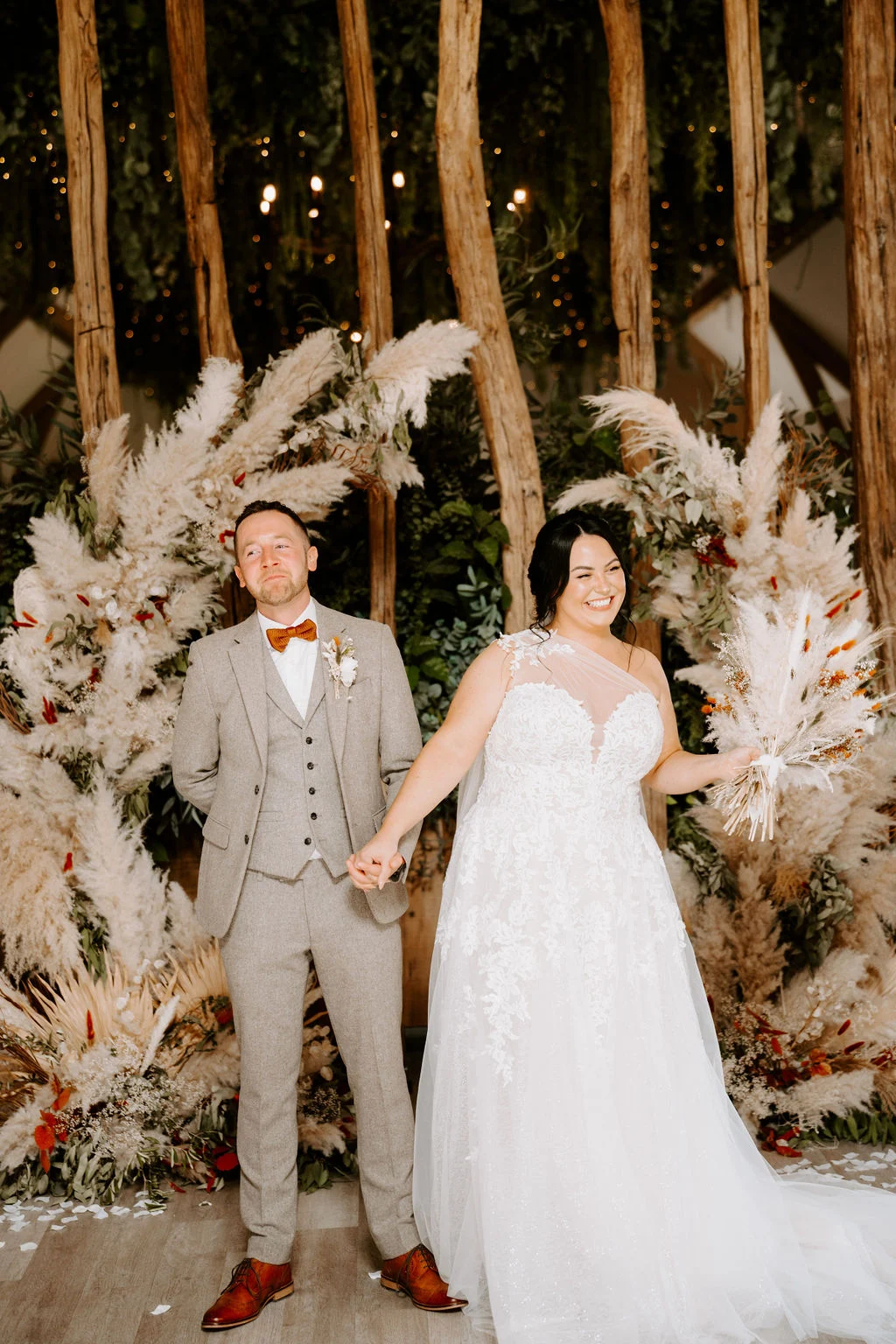 Hannah and Tom holding hands beside their floral ceremony backdrop at The River Barn