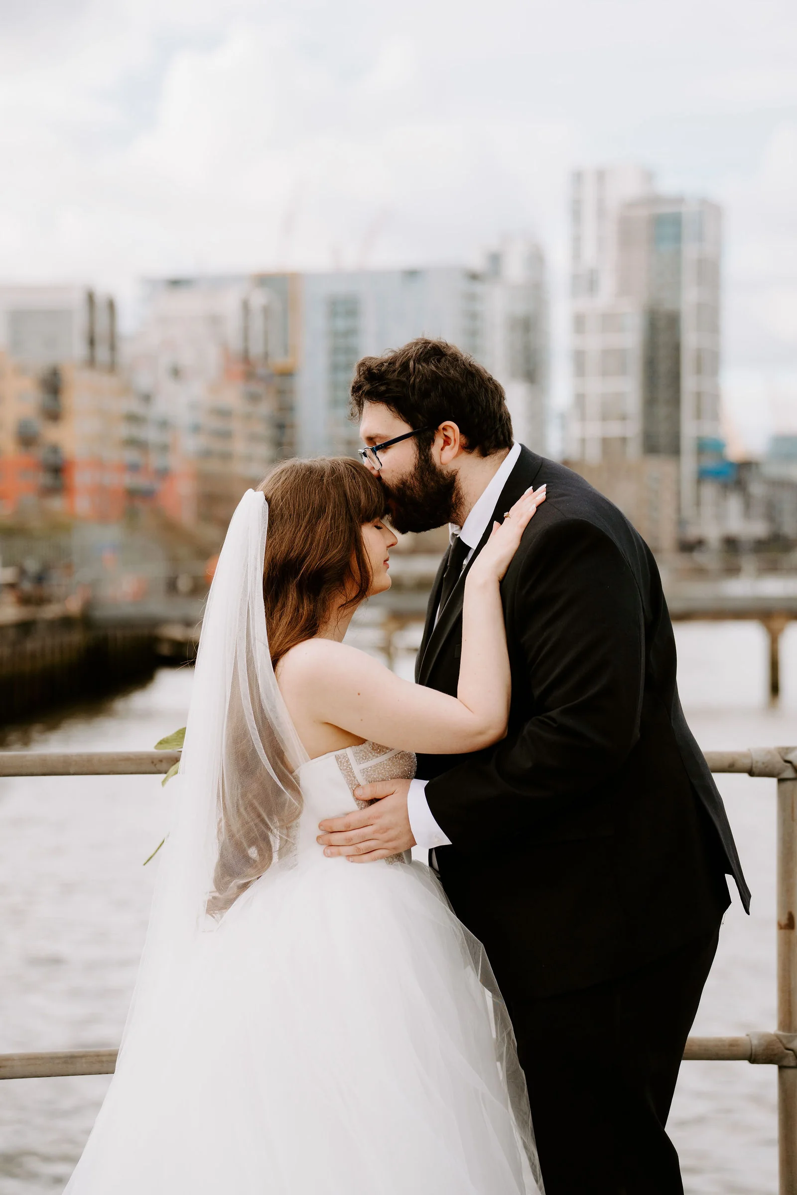 A groom in glasses and a black suit kisses the forehead of his bride in a veil by a river railing, with apartment towers across the water