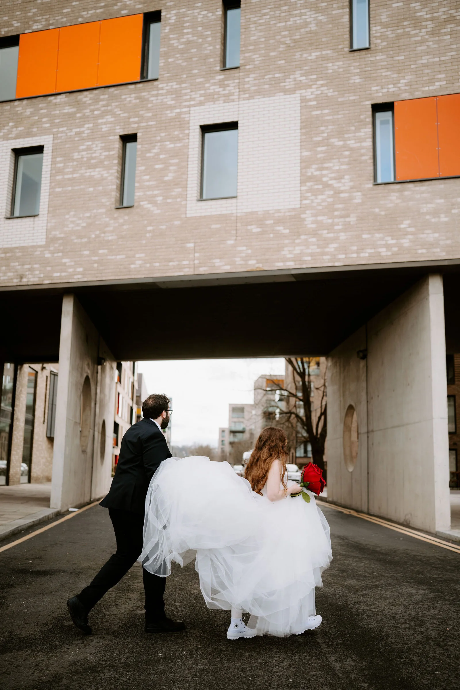 A groom gathers up the tulle skirt of his bride, who carries a bouquet of red roses, as they walk under a brick building with orange panels