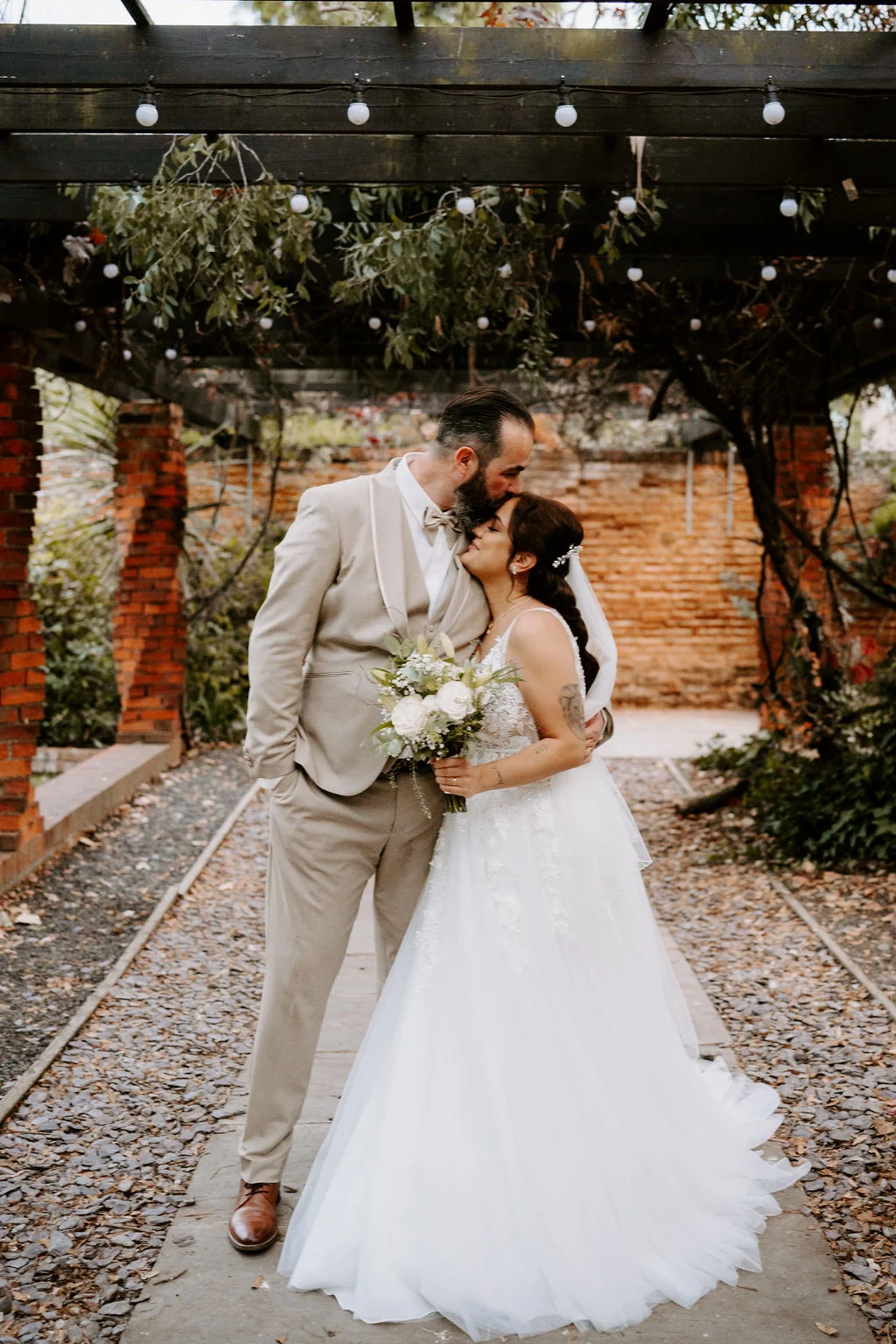 Hugo and Mel embracing with her bouquet in the gardens.