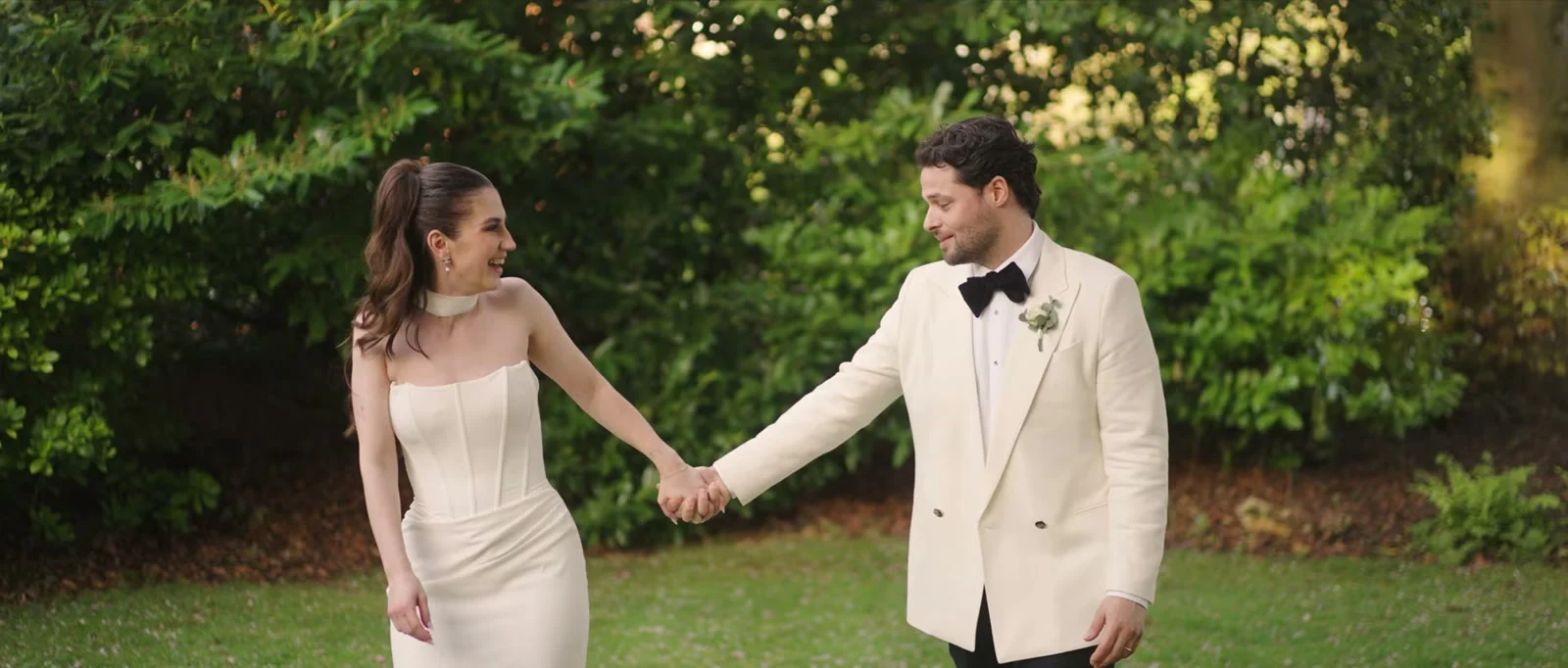 A laughing bride in a strapless white dress looks back at her groom in an ivory dinner jacket and black bow tie as they walk hand in hand across a lawn with dense green shrubs behind them