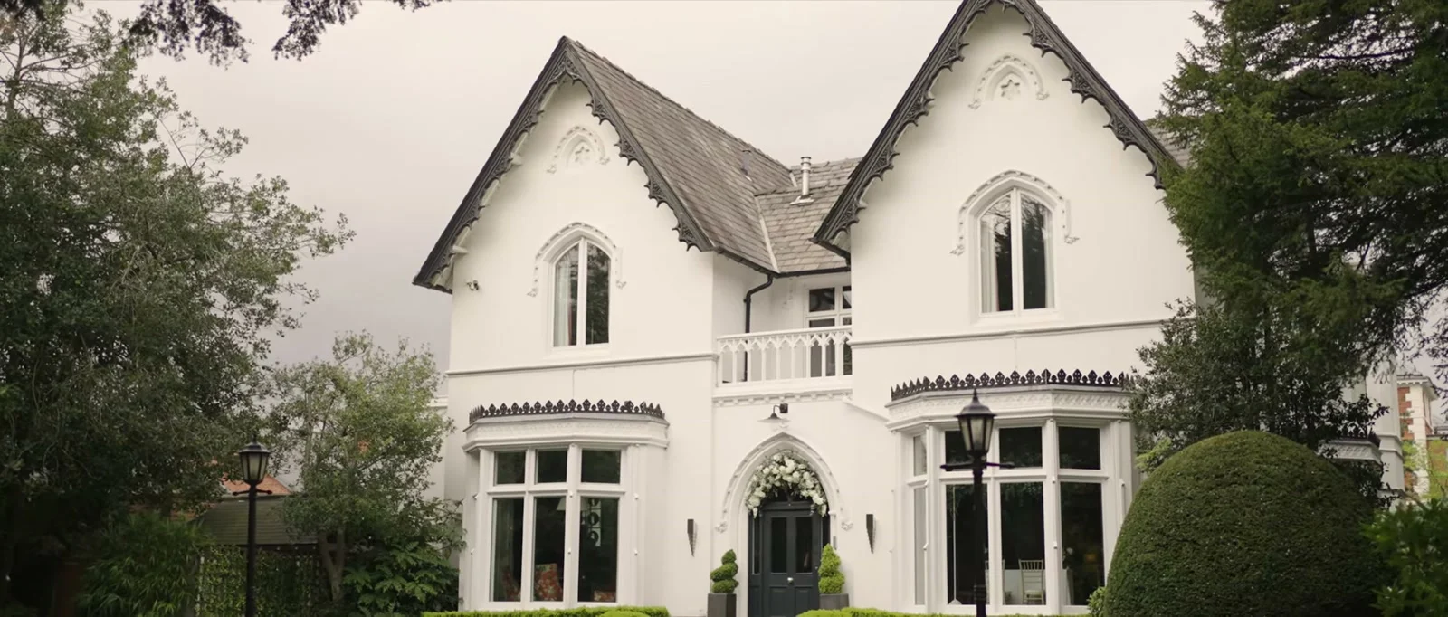 The white Victorian front of Didsbury House Hotel with pointed gables, dark decorated bargeboards, bay windows and white flowers over the arched front door