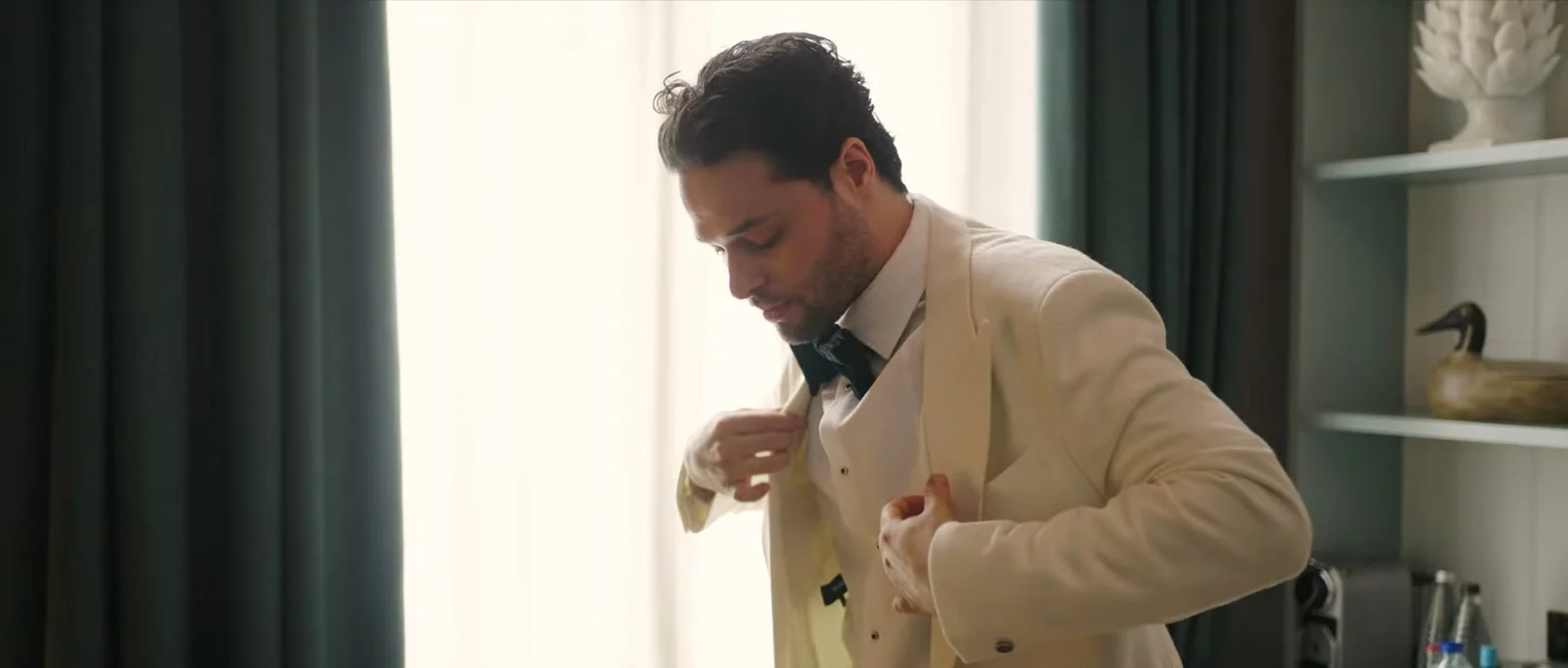 A groom in an ivory jacket looks down as he adjusts it over his dark bow tie beside a tall window with dark green curtains